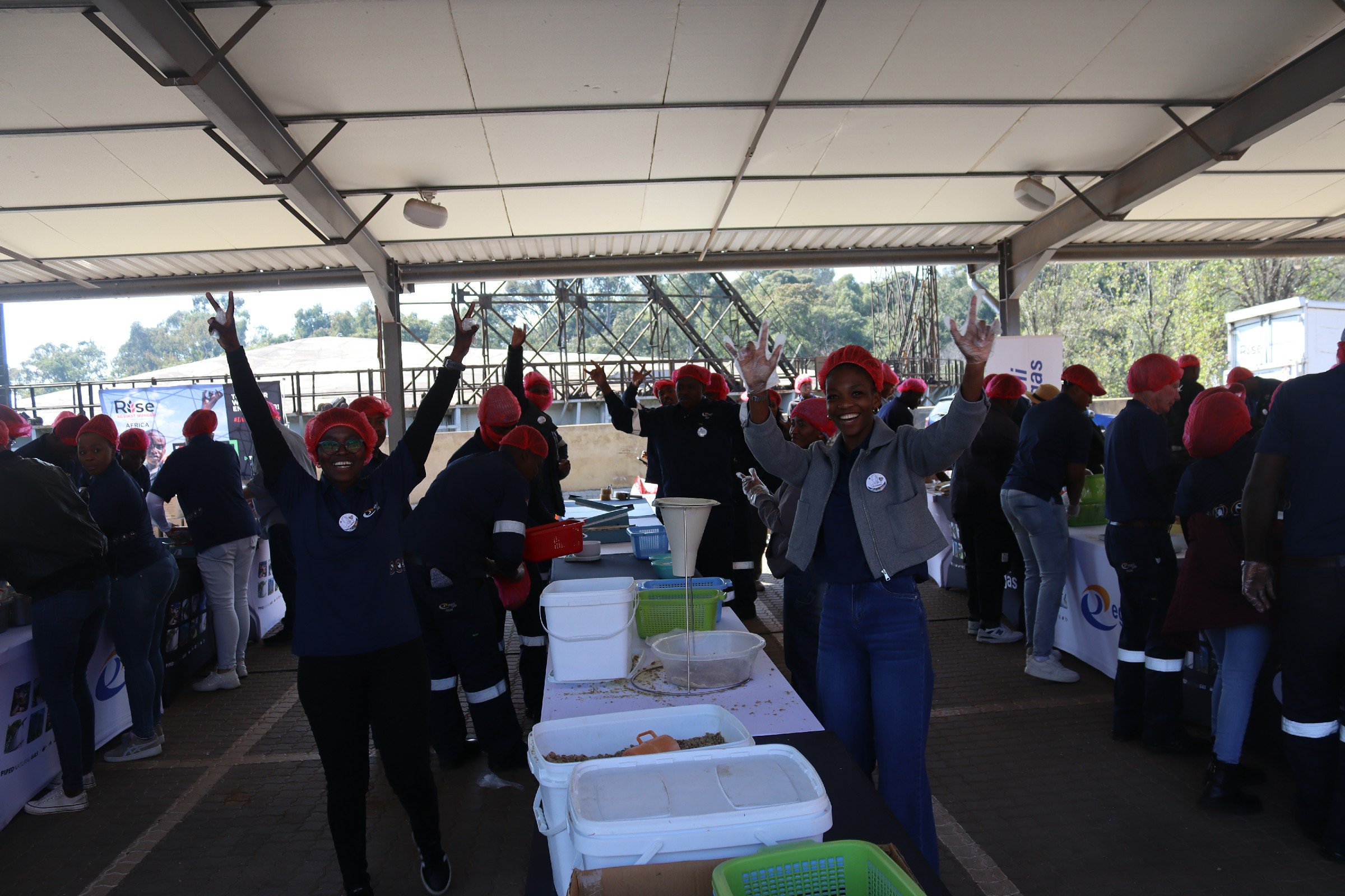 People wearing red hairnets and dark uniforms working at a table with containers and food items outdoors, under a large covered area, smiling and making peace signs.