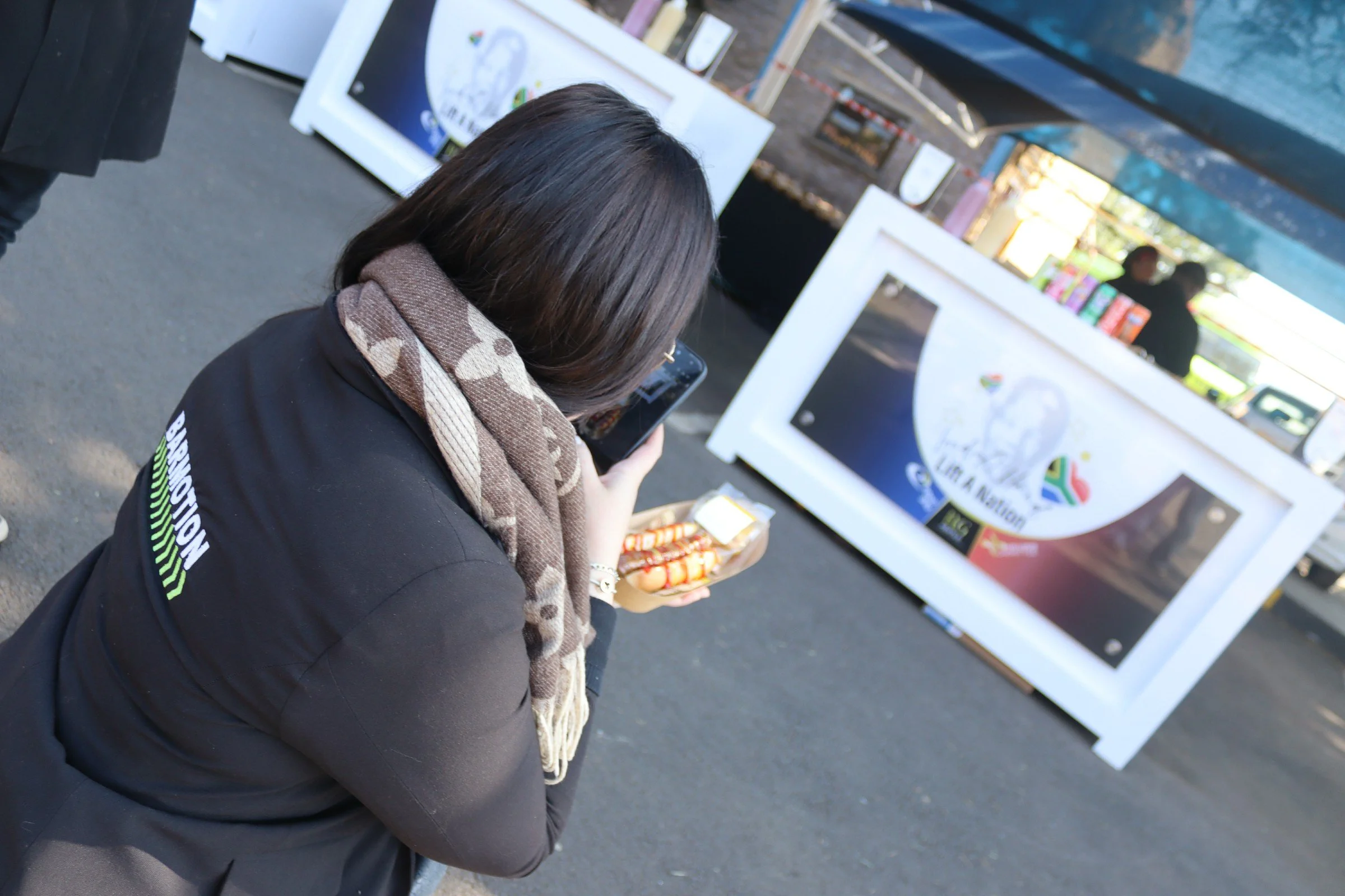 A woman with dark hair, wearing a black jacket and a brown patterned scarf, is standing outdoors looking at her phone. Behind her are display boards with graphics and a sign with the South African flag and text.