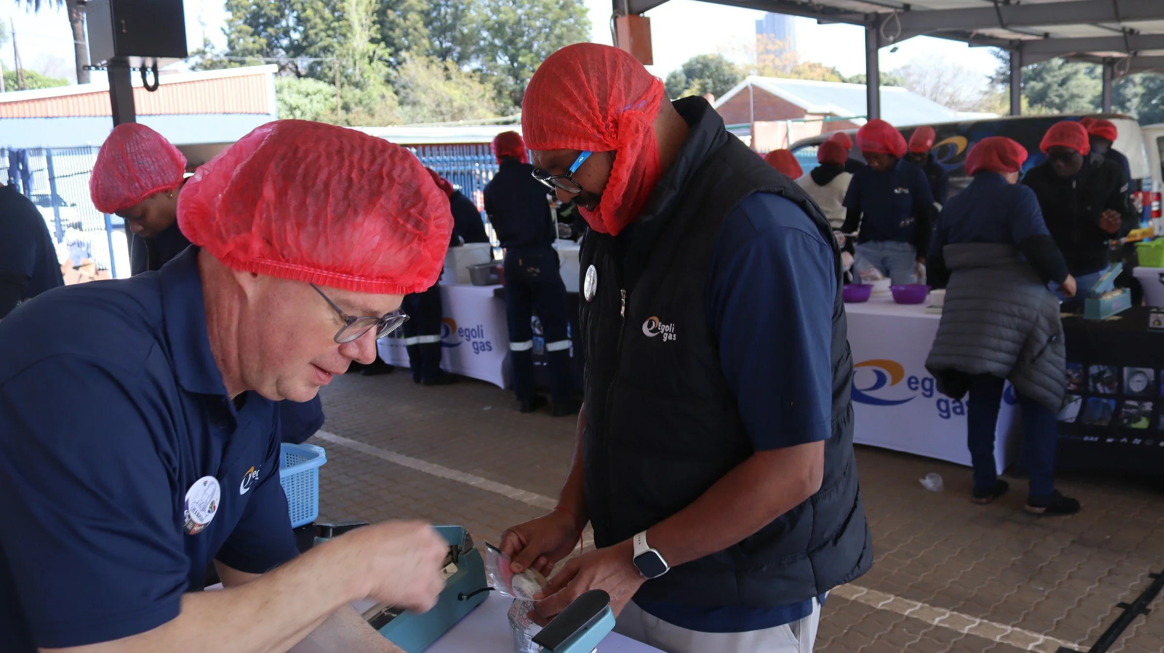 Two men wearing red hairnets and dark vests are engaged in a transaction at an outdoor event. Background shows multiple people in similar attire, tables with banners, and trees in the distance.