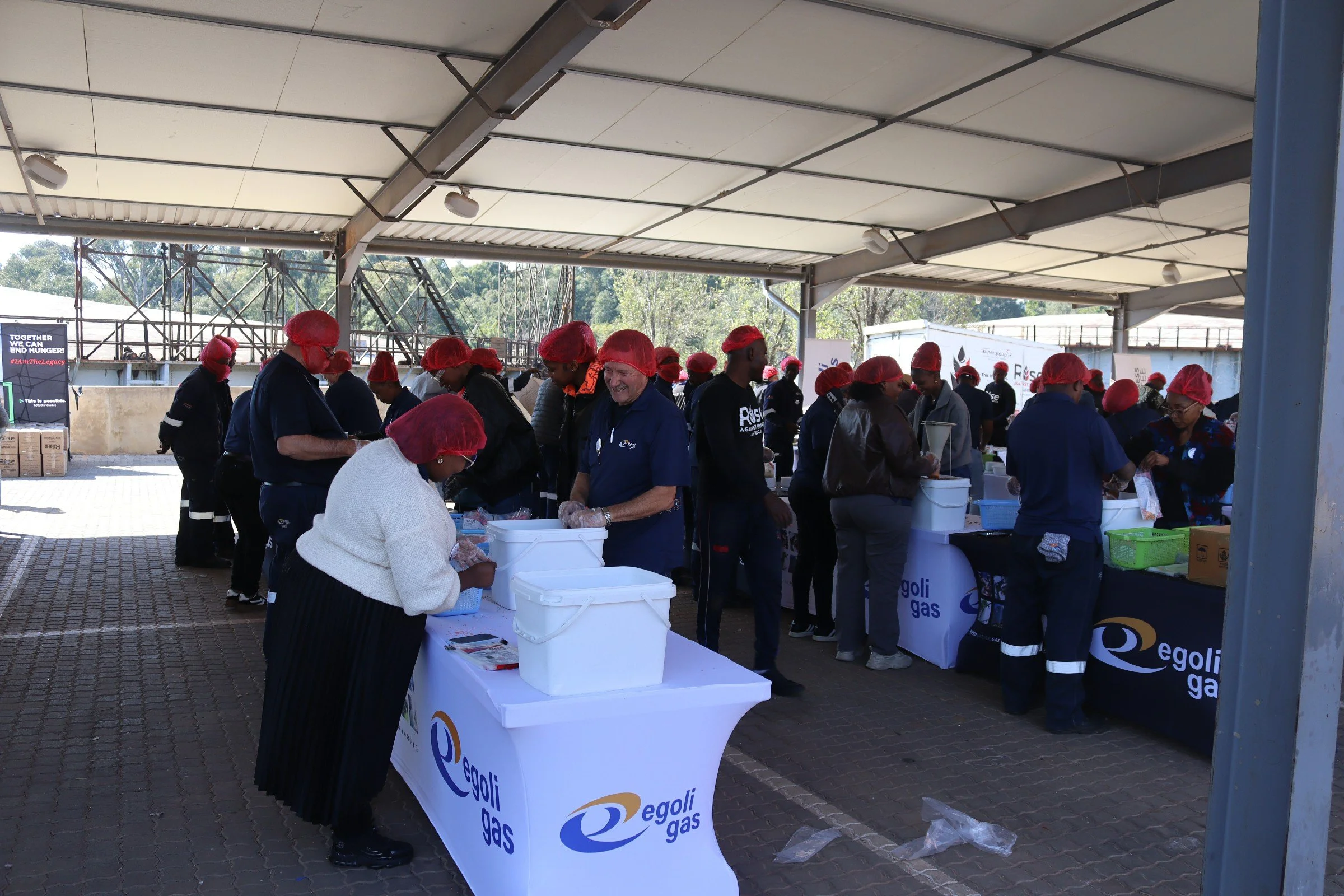 People wearing red hair covers serving food or distributing items at a community event under a large open-air shelter, with their focus on a table with white containers and promotional banners for Egoli Gas.
