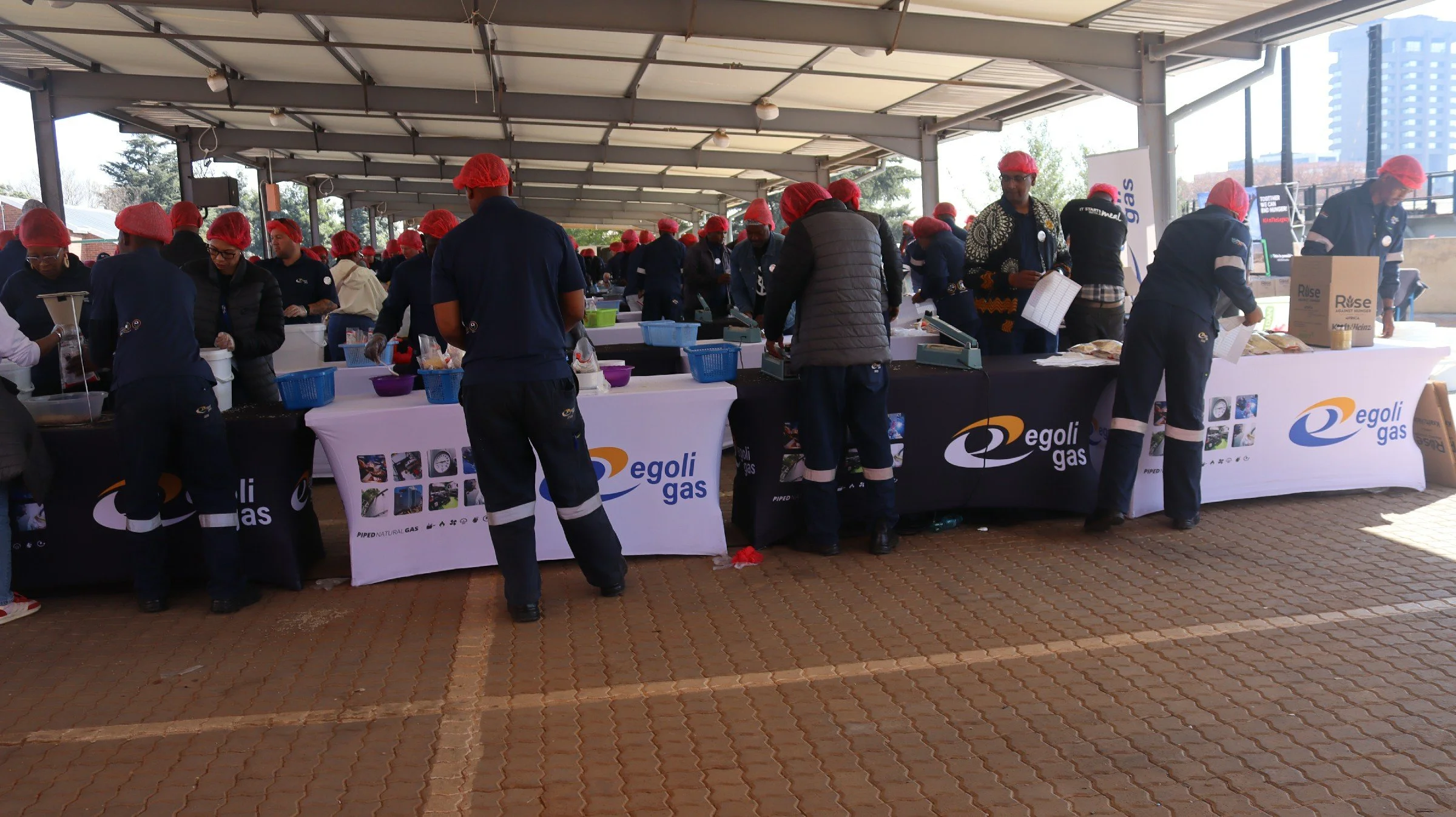 Group of people wearing red hairnets working at tables covered with Egoli Gas banners under a pavilion with a cityscape background.