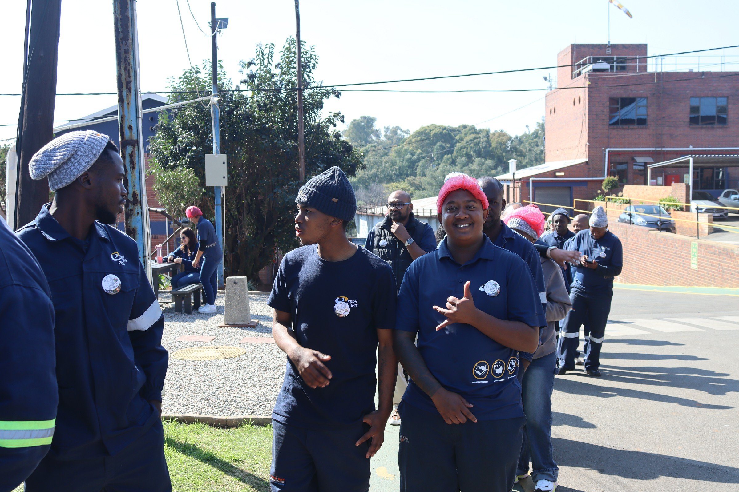 Group of people standing outdoors, engaged in conversation. Some individuals are wearing navy uniforms, and a woman is smiling at the camera with a pink hair cover. The background features trees, a brick building, and parked cars.