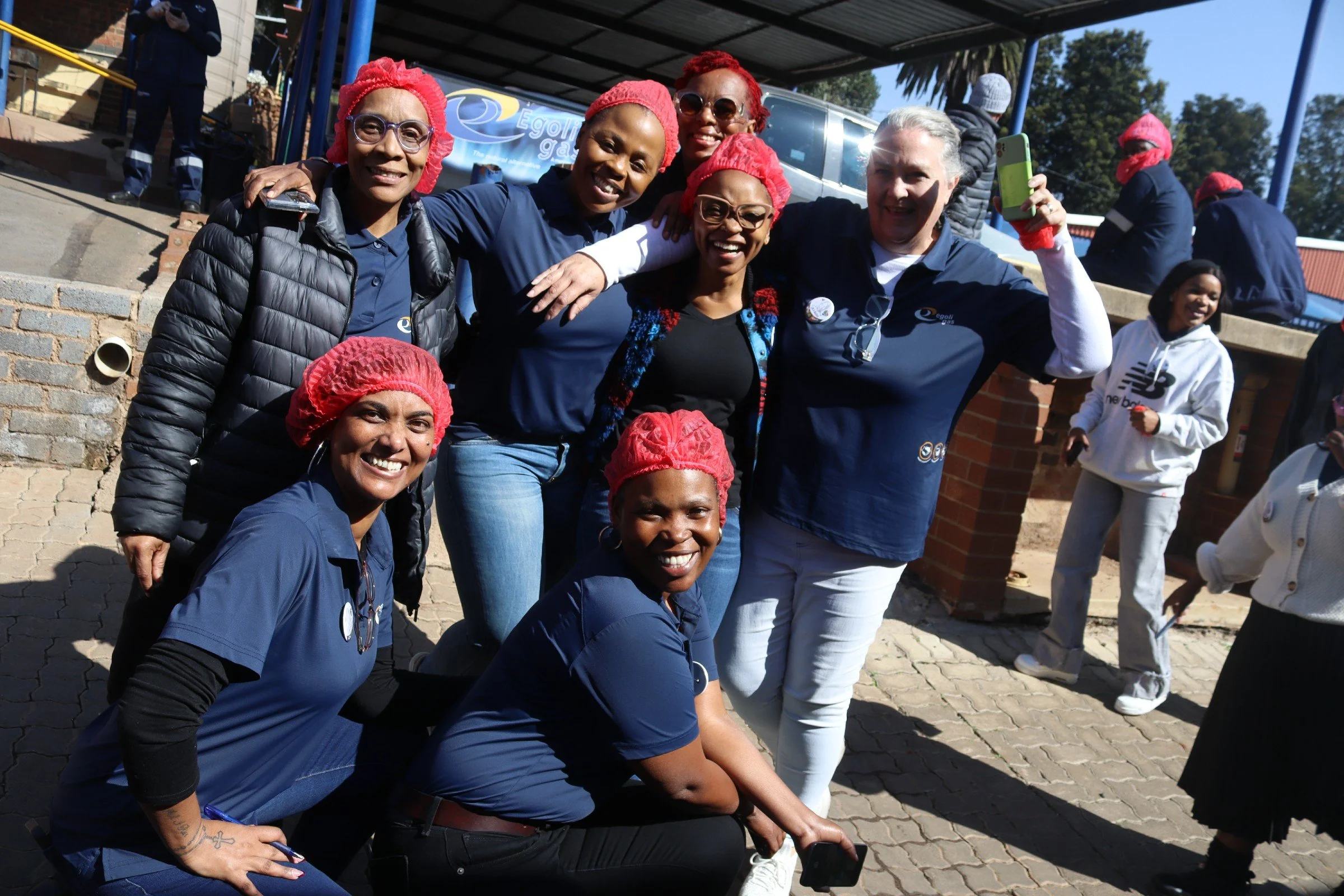 Group of women smiling and posing for a photo outdoors during a gathering or event, some wearing red hair coverings.