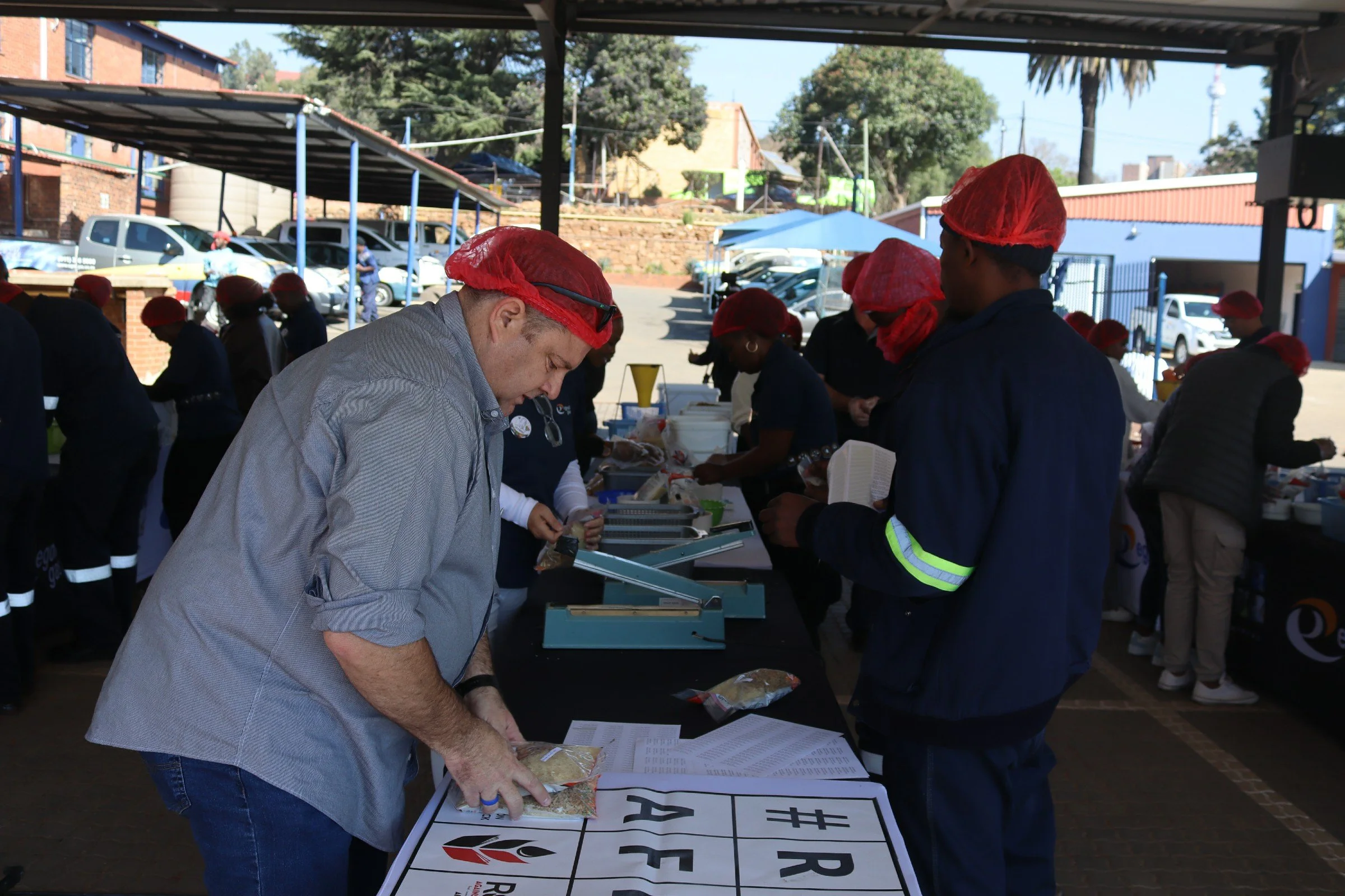 People wearing red hairnets and dark clothing gather at tables outdoors, sorting packages of food or supplies under a shelter with parked cars and trees in the background.