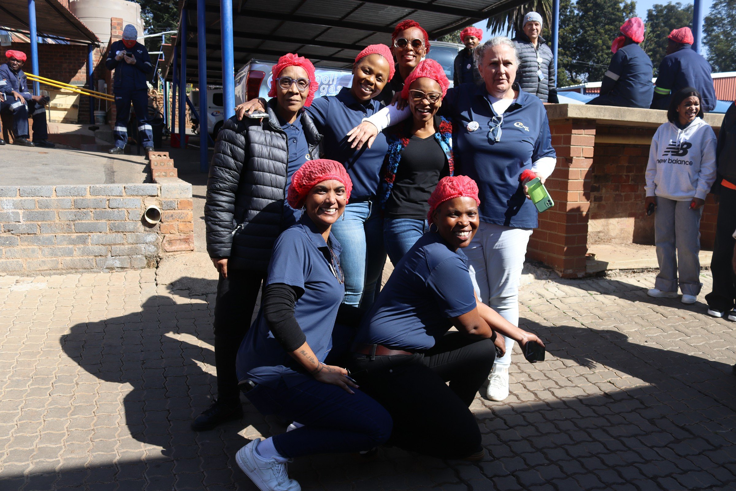 A group of nine diverse women wearing casual clothing, some with red hairnets, gathered outdoors in bright sunlight, smiling and posing for a photo, with a brick and metal structure and other people in the background.