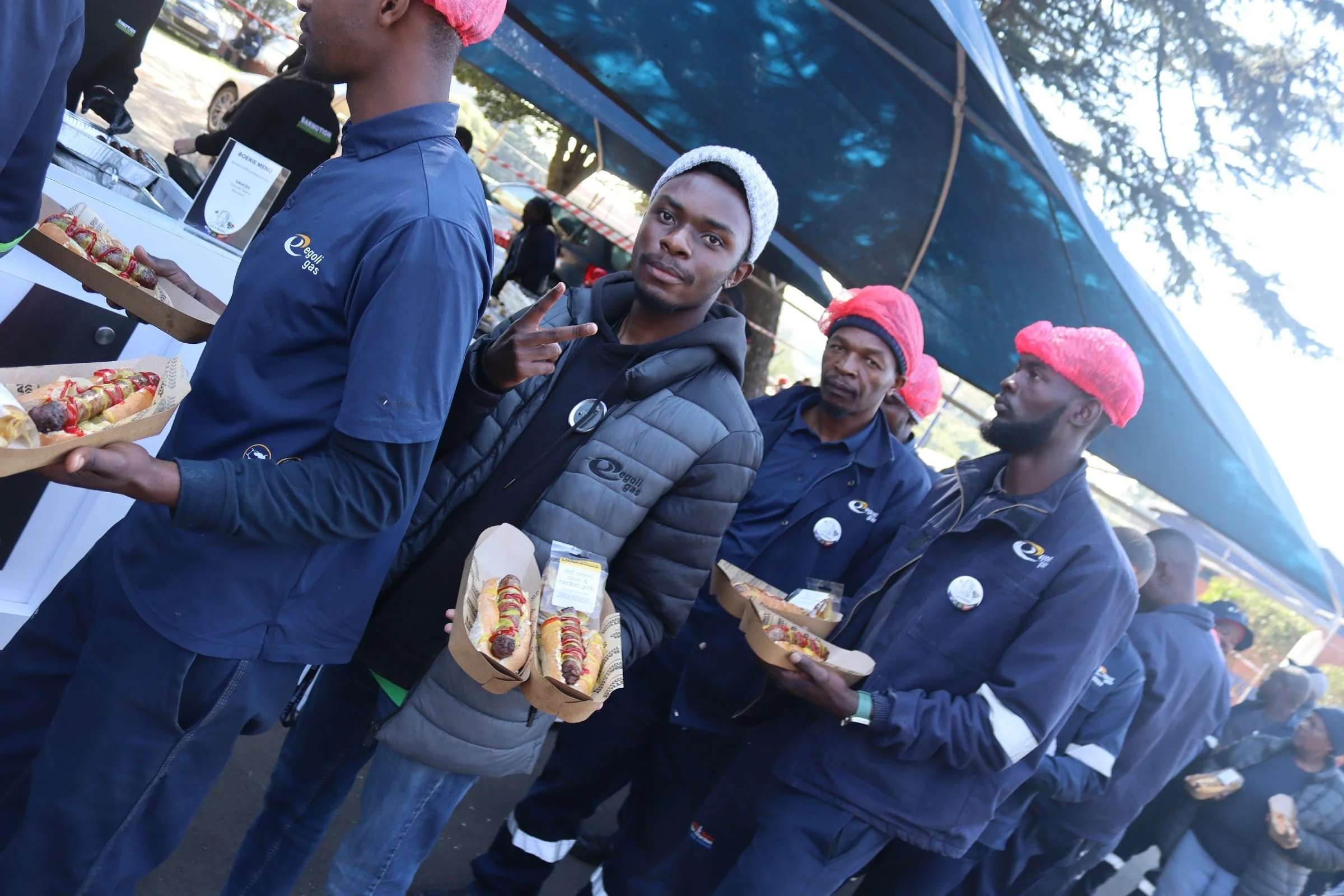 Young man in a black jacket and gray vest making a peace sign while holding a tray of hot dogs at an outdoor event, with other people in the background under a blue canopy.