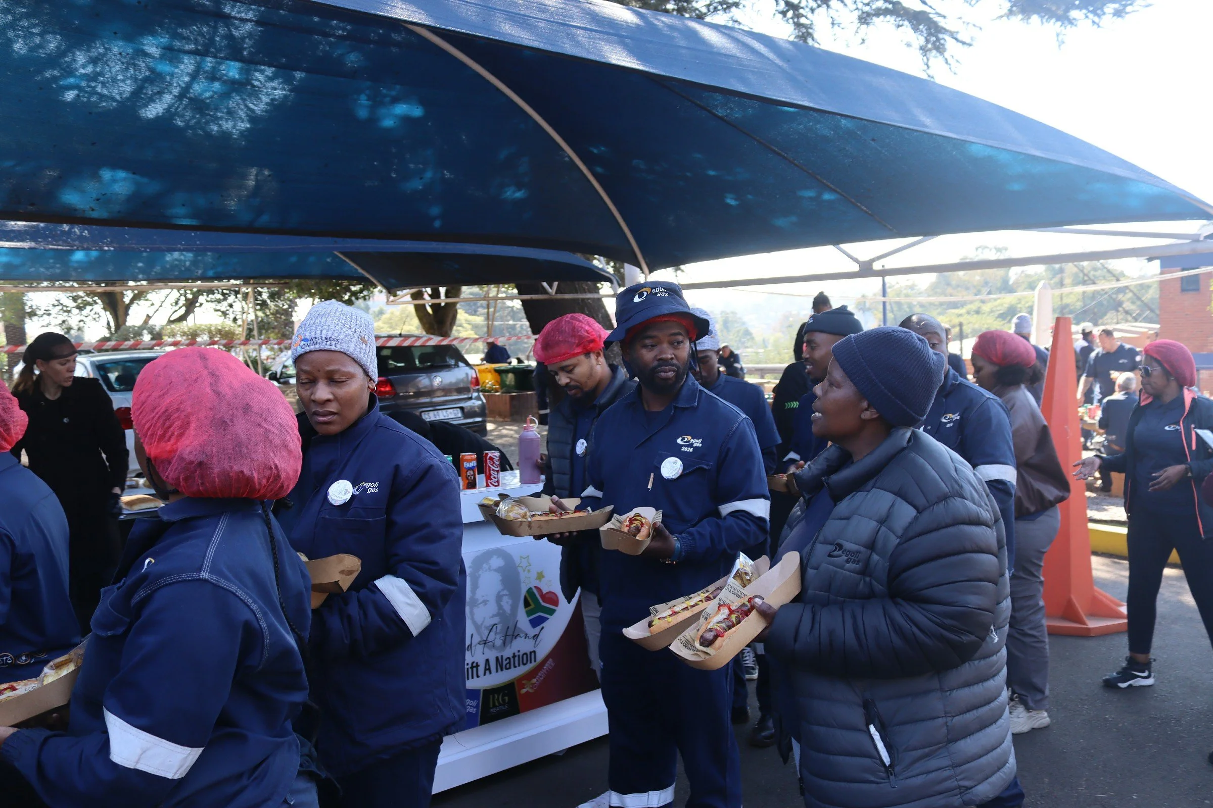 People wearing jackets and hairnets standing in line at an outdoor food distribution event, with cars and trees in the background.