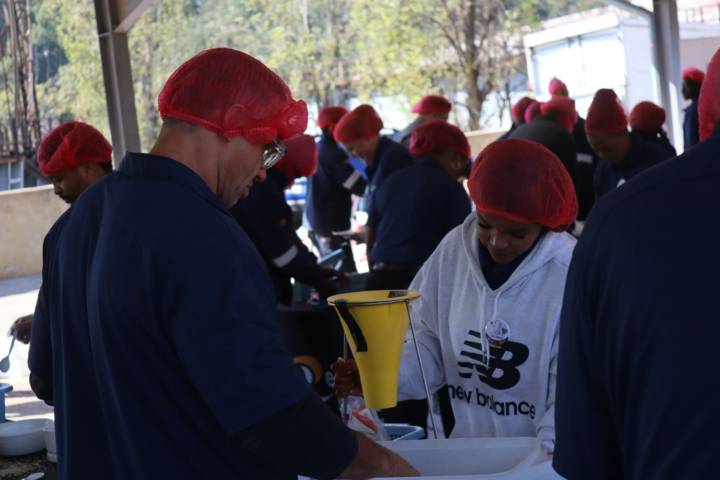 A group of people wearing red hairnets and dark clothing gather around a table outdoors, preparing or processing food under a shelter with trees in the background.