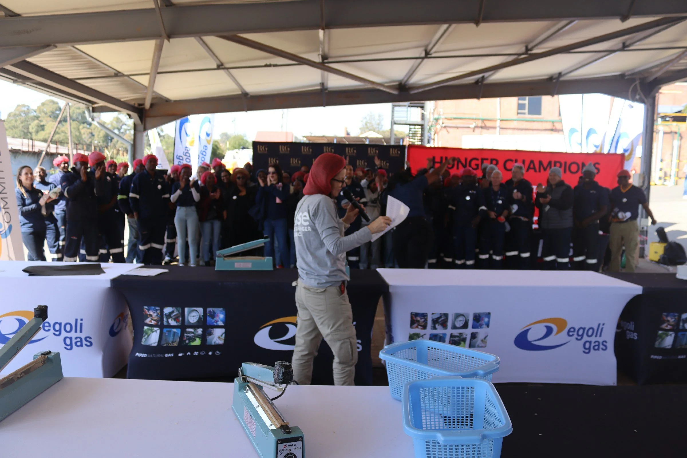 A group of people wearing navy blue uniforms and pink hair coverings gathered under a tent, raising their hands in a celebratory manner. A woman in the foreground is speaking into a microphone. There is a red banner with the text 'HUNGER CHAMPIONS' i