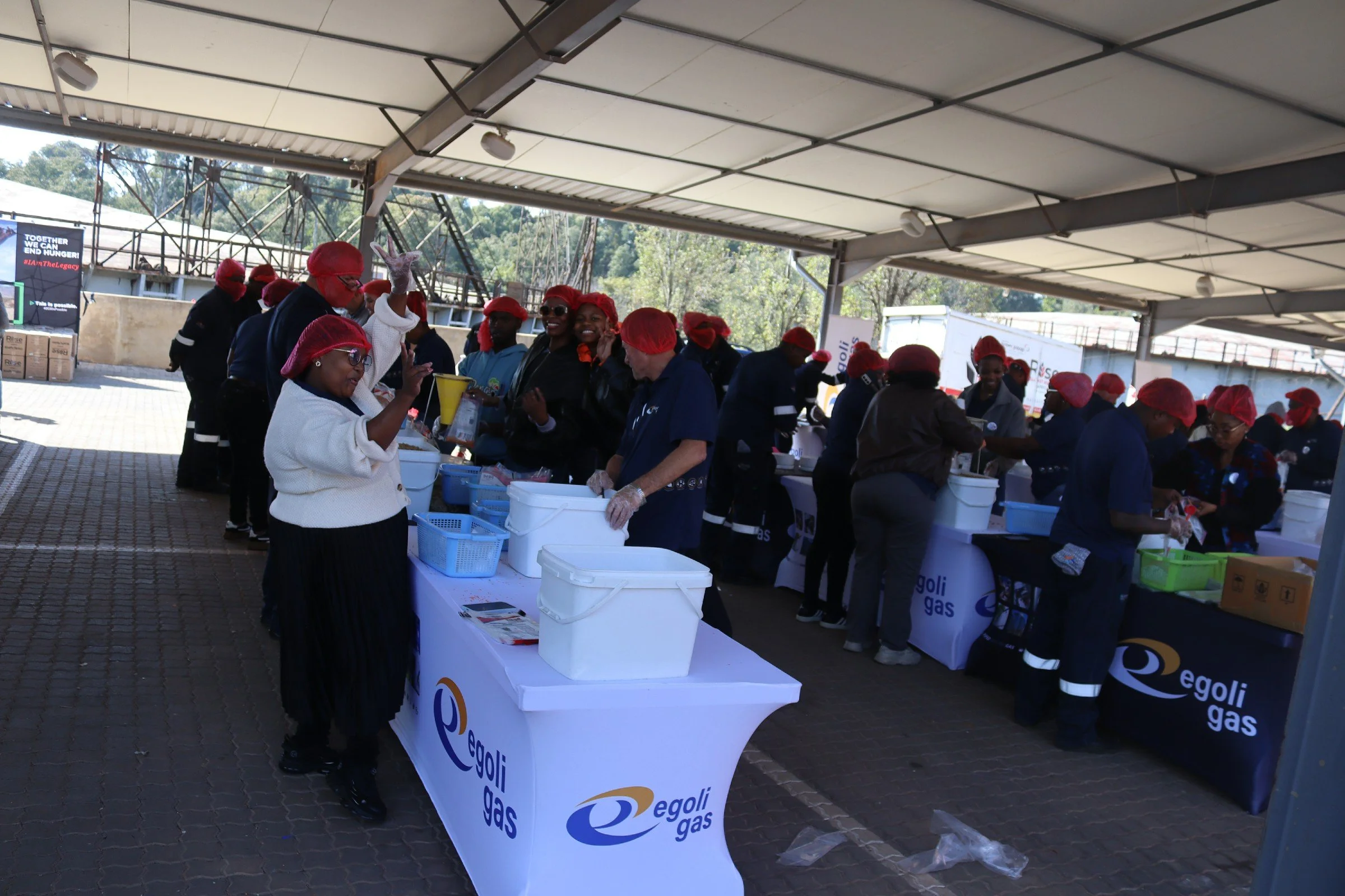 Outdoor event with a large group of people wearing red hair coverings and dark clothing, serving food or drinks at tables branded with Egoli Gas logos under a canopy.
