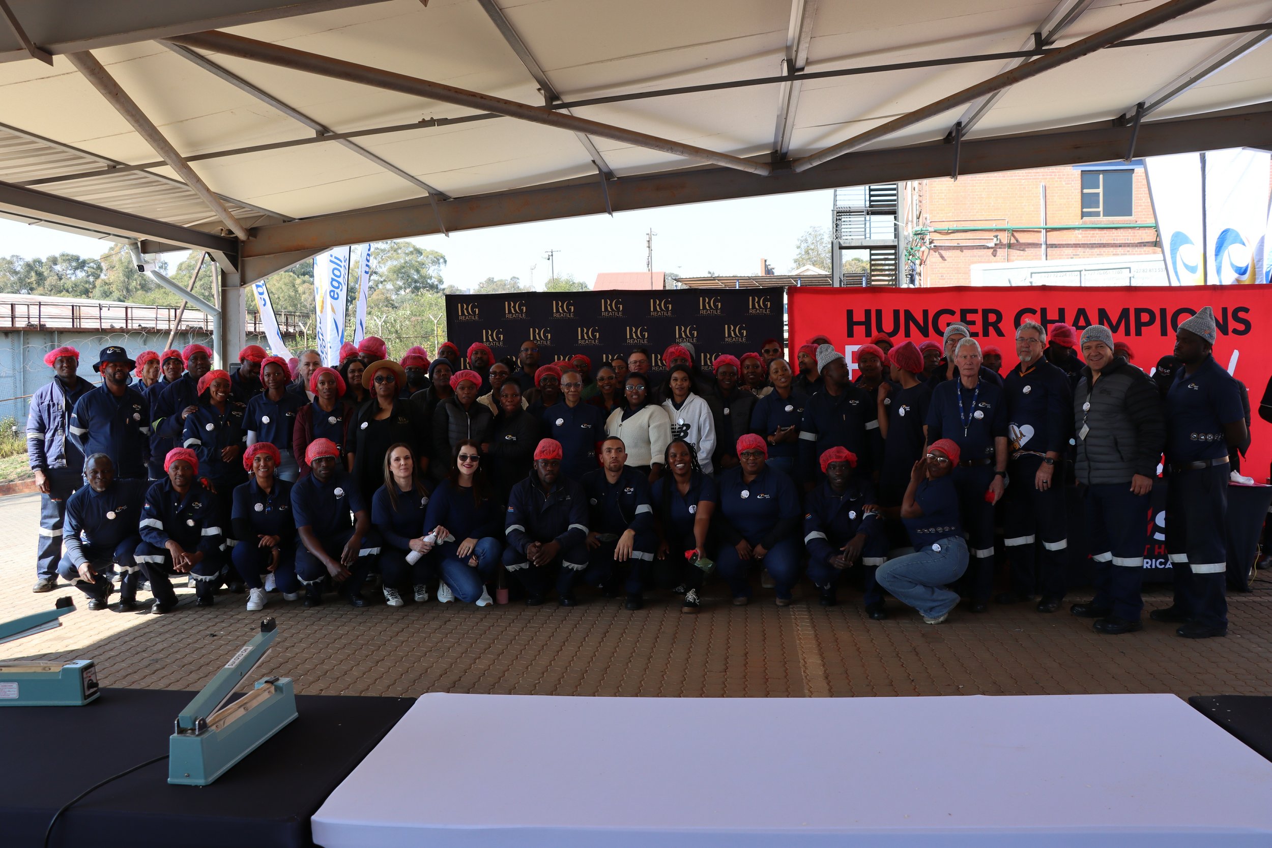 A large group of people wearing matching navy blue uniforms and red or gray hairnets, standing under a canopy with a sign that reads 'HUNGER CHAMPIONS' in the background. Some people are smiling and posing for the photo.