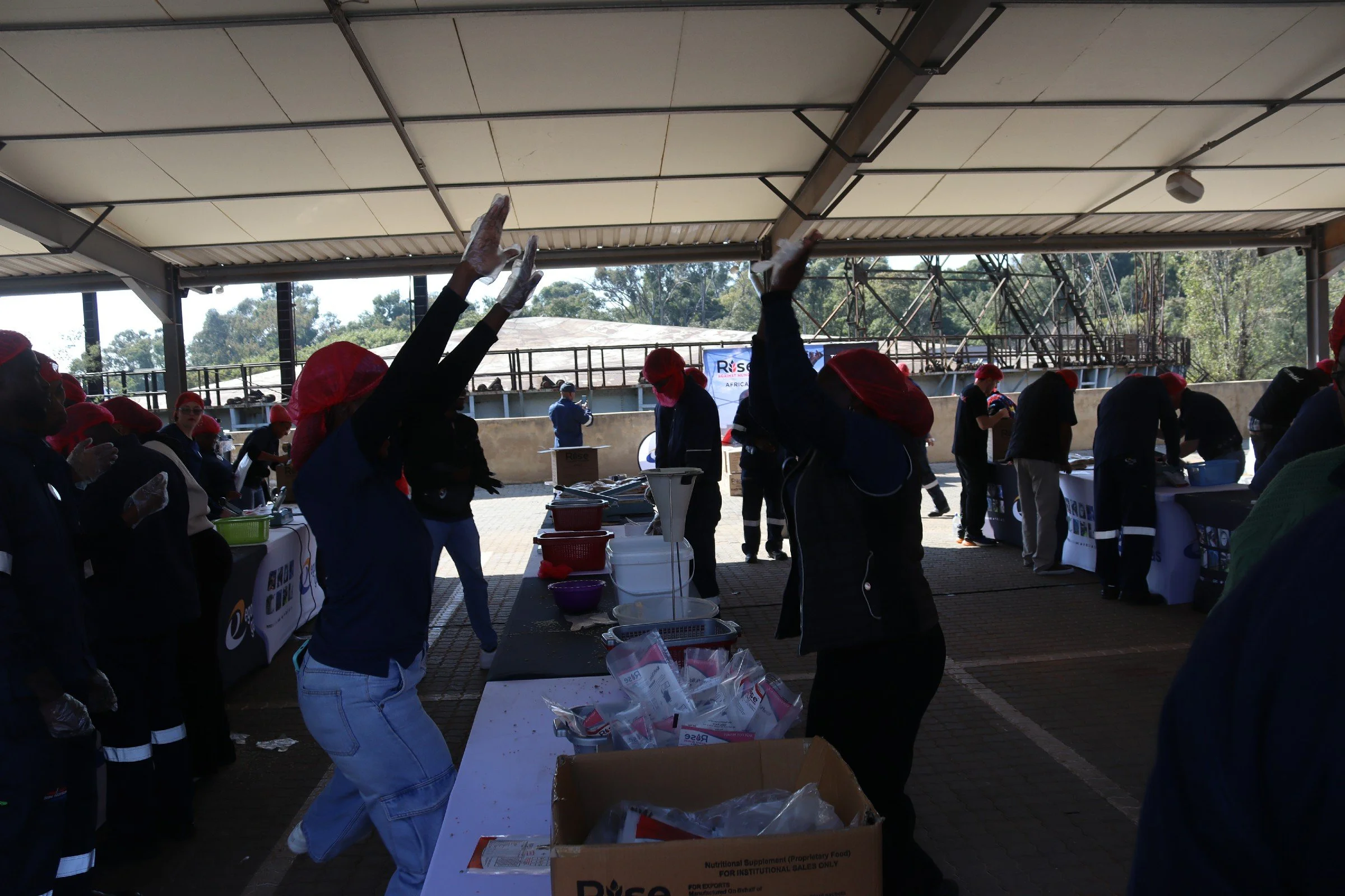 Group of people working at a food distribution event outdoors under a canopy, with tables, boxes, and supplies, some raising their hands.
