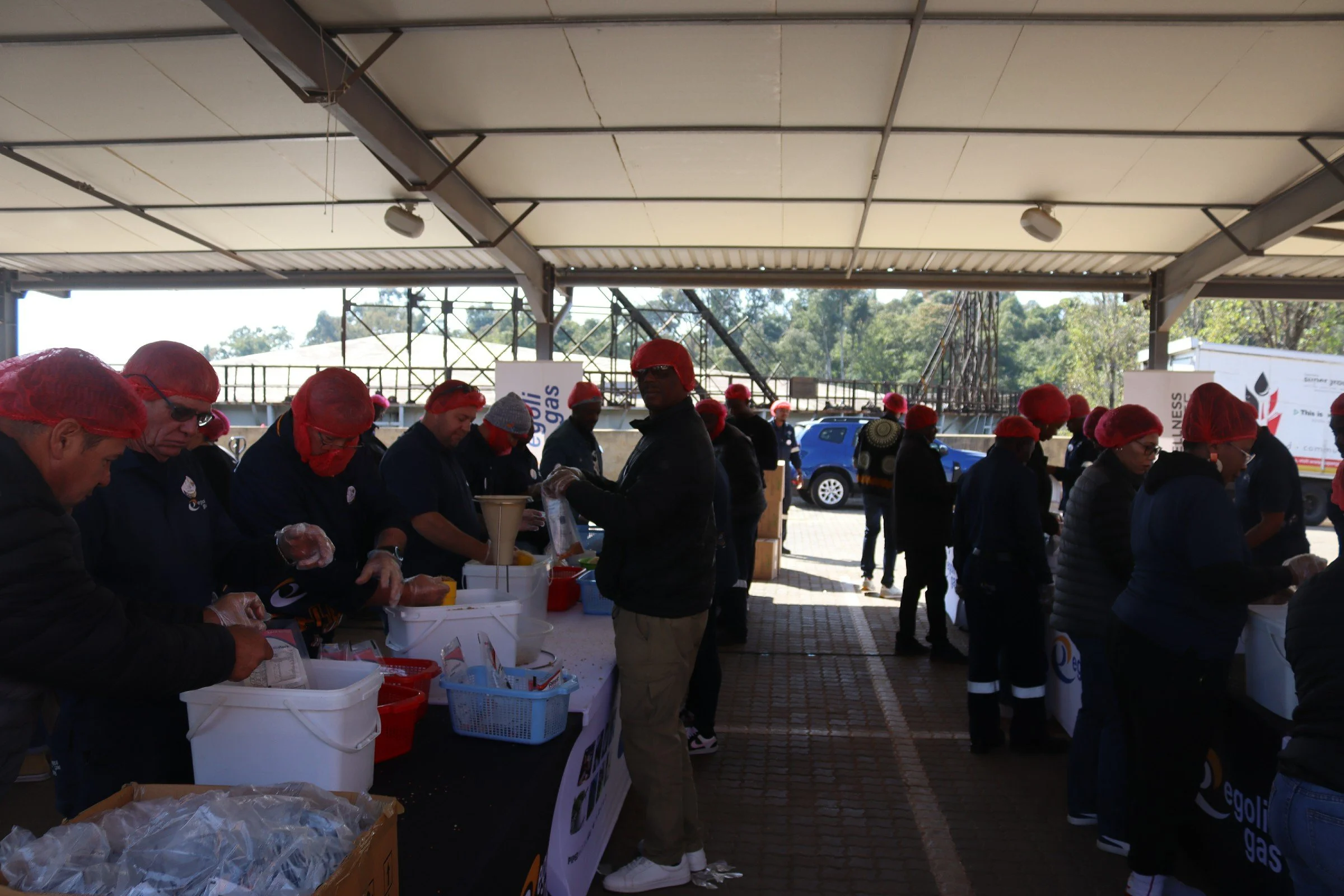 People wearing red hairnets and gloves, standing in line at a food distribution event outdoors under a shelter, with carts of supplies and a sign in the background.