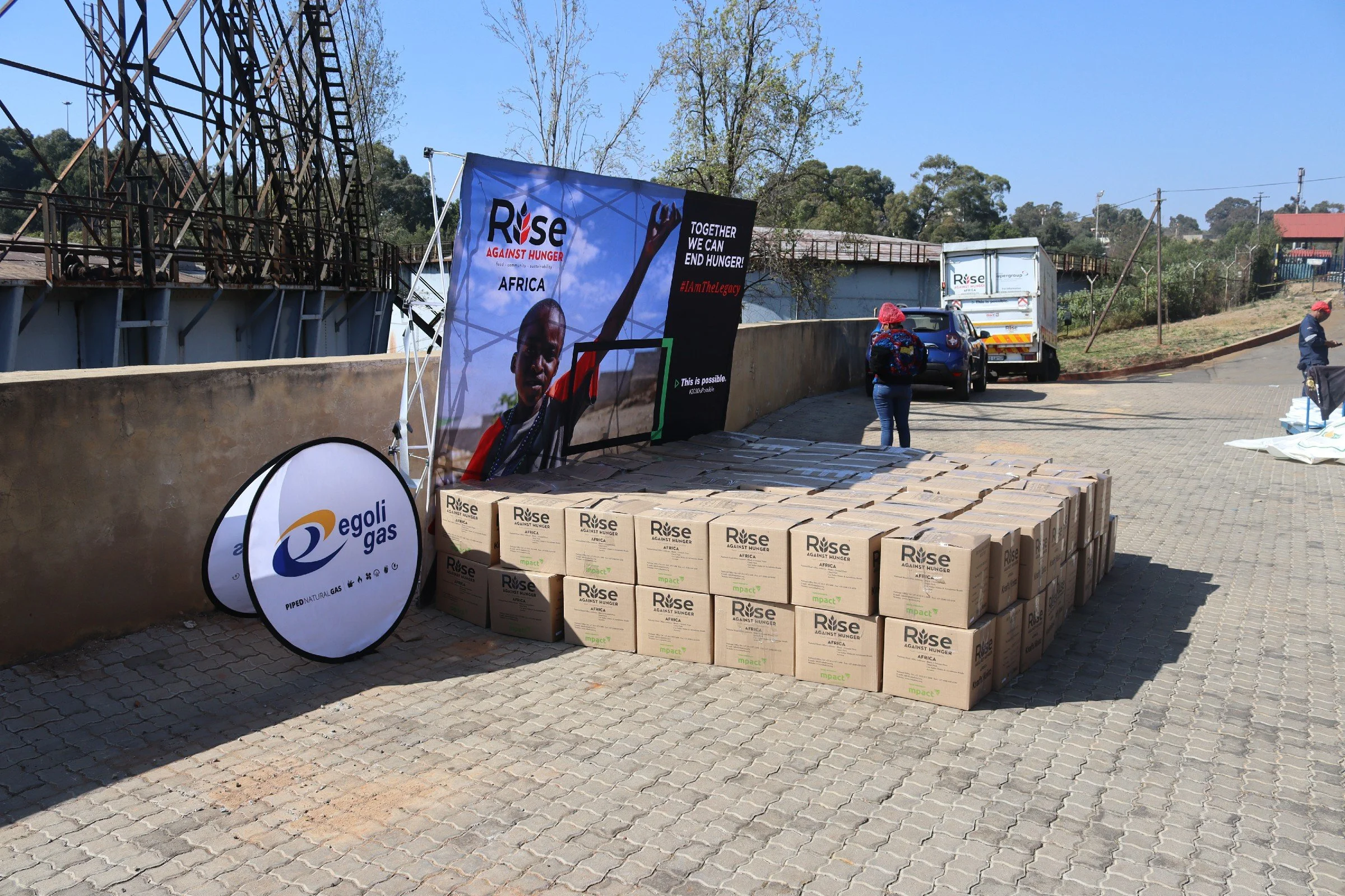 Streetside setup with campaign banners and boxes of aid supplies for an hunger relief event in Africa. Two people are seen working, with a truck and parked vehicle in background.