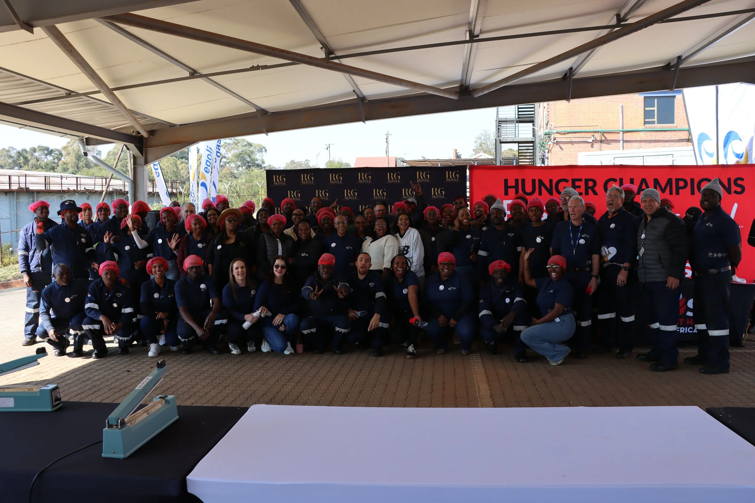 A large group of people gathered under a tent at a charity event for Hunger Champions, wearing red hairnets and standing in front of a red banner with black text. There are tables with equipment in the foreground and banners with logos in the backgro