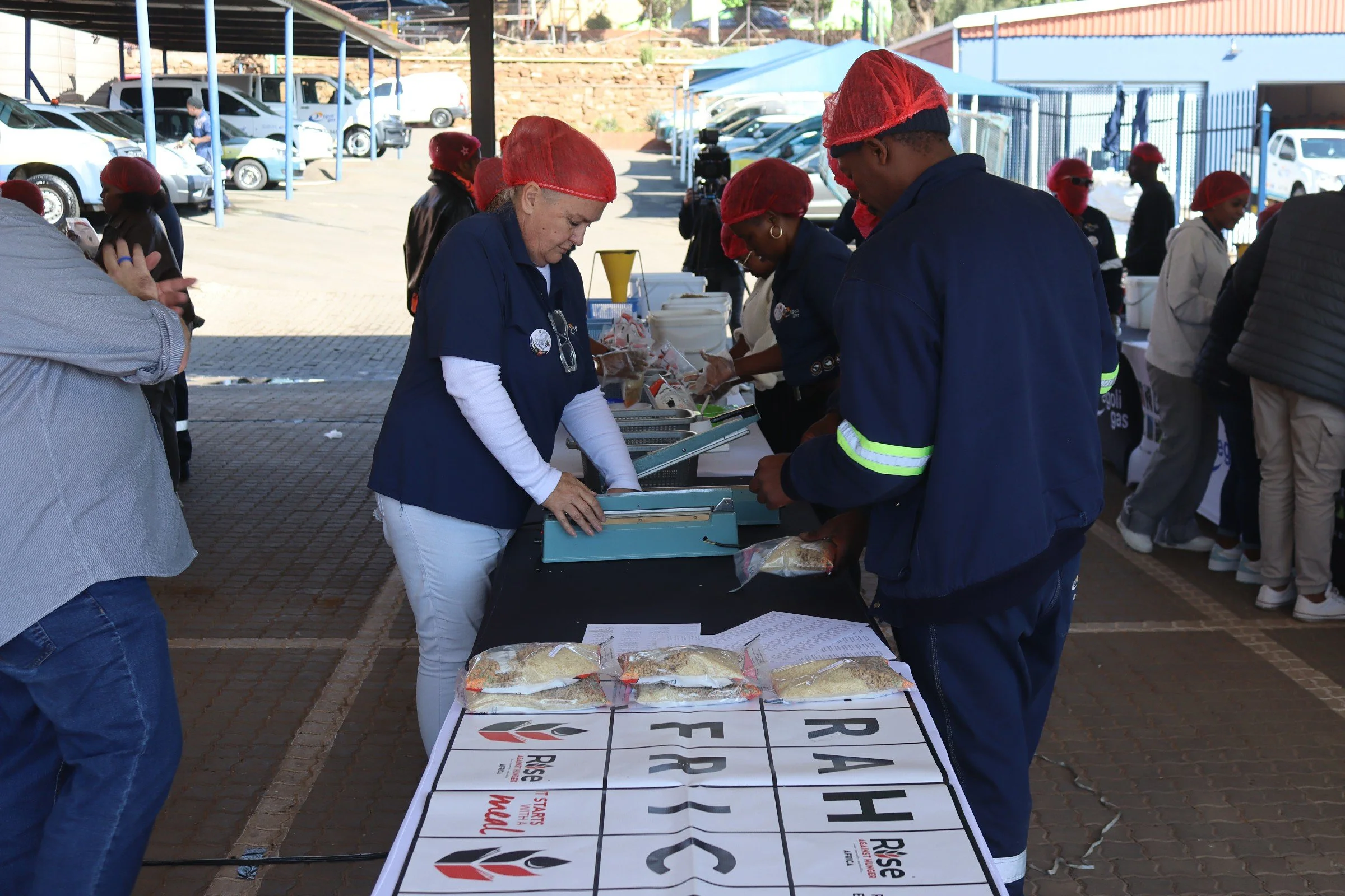 People wearing red hairnets and navy uniforms gather around tables with packaged food, outdoors at an event with parked cars and tents in the background.