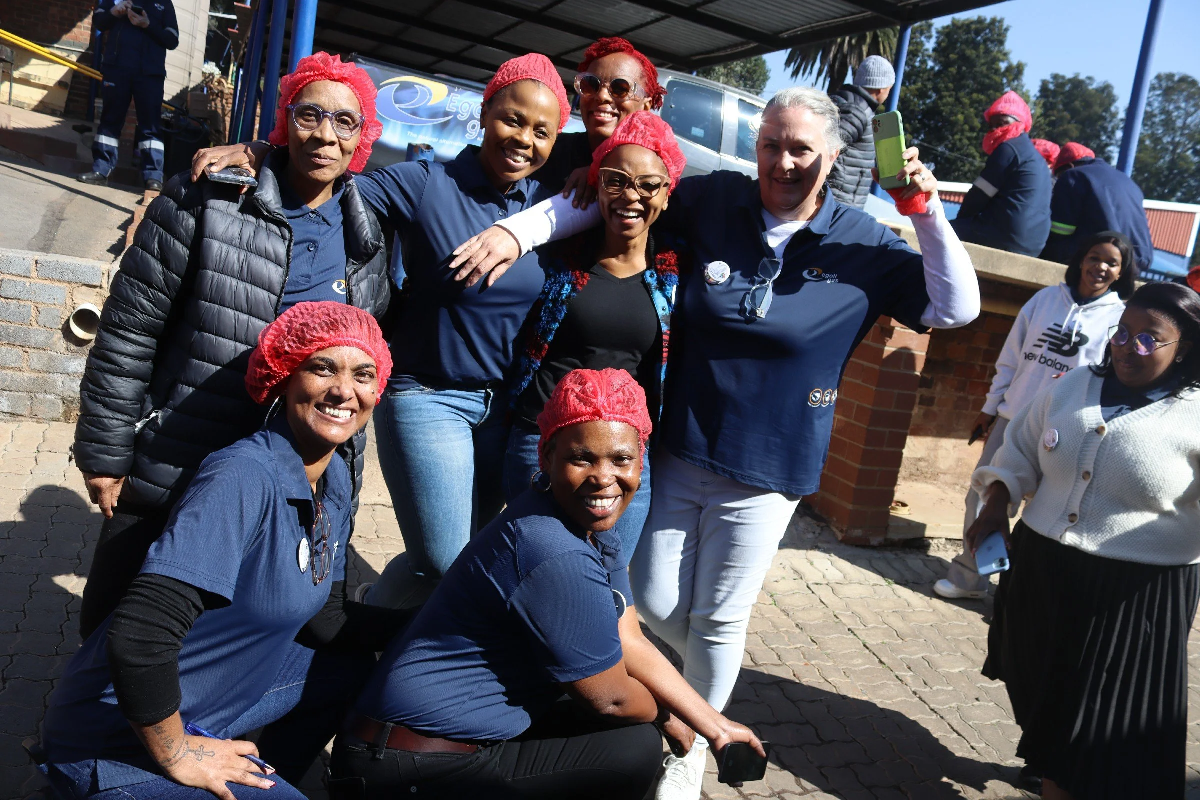 Group of smiling women, some wearing red hairnets, gathered outdoors for a photo with arms around each other. They are dressed in casual and workwear, with a few holding smartphones.