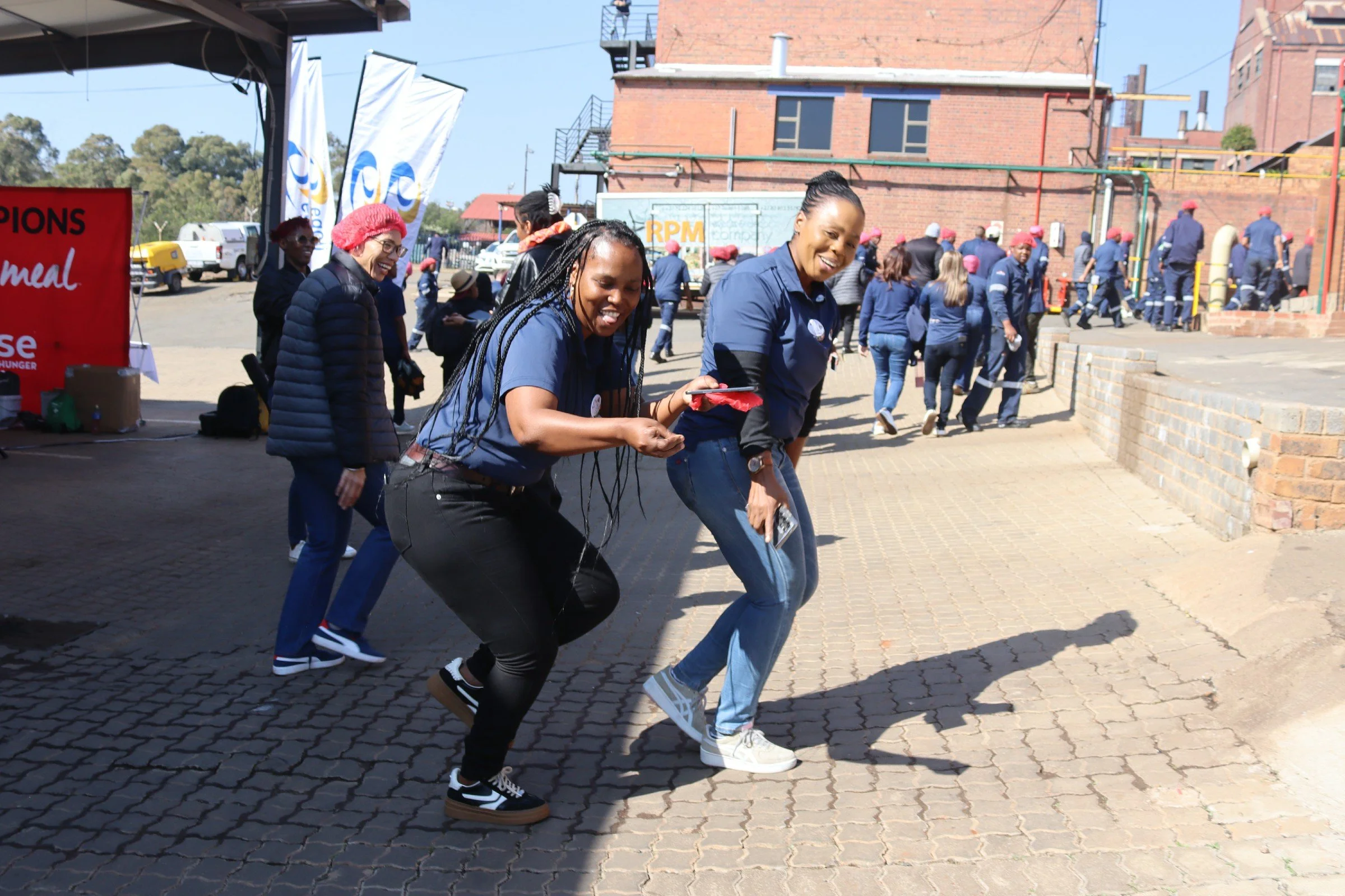 Group of people dancing outdoors on a cobblestone area with brick buildings in the background. Many are wearing blue shirts and some are wearing red hats.