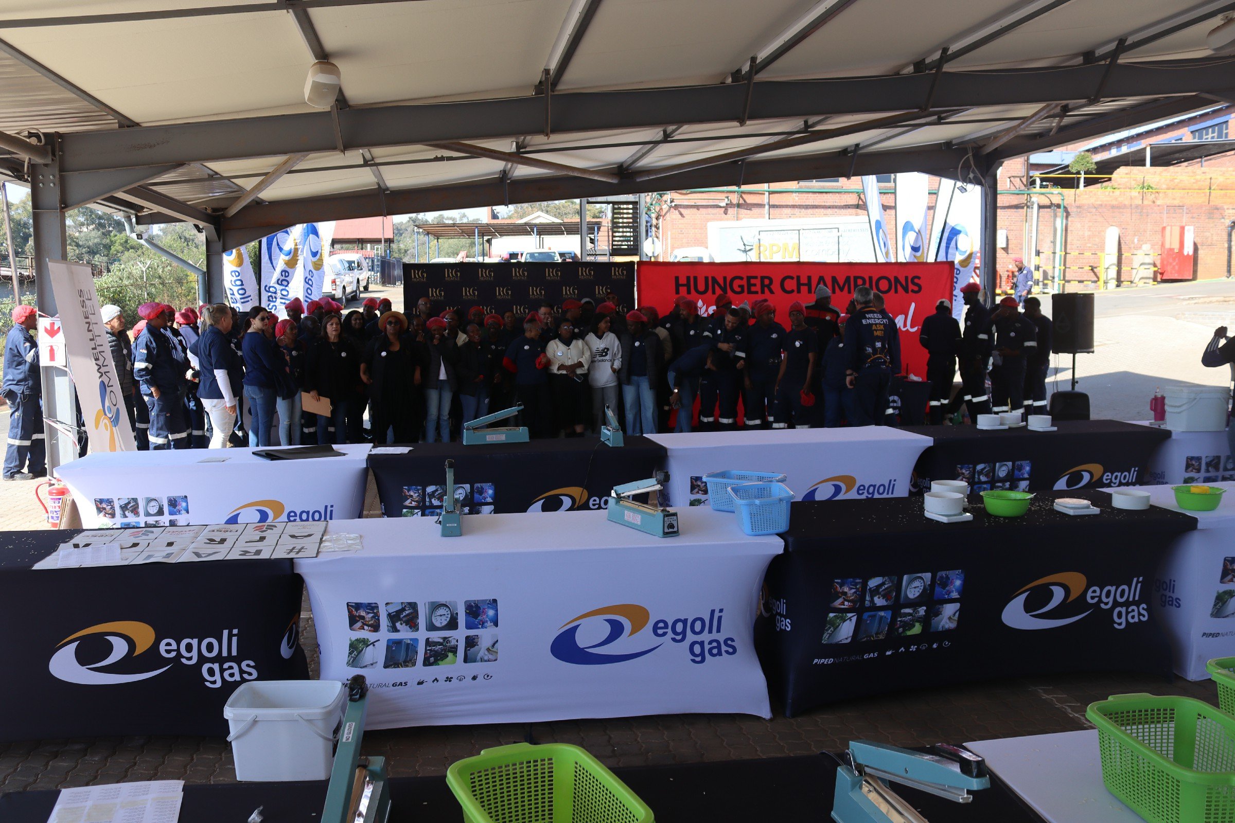 A group of people wearing red hats and dark clothing gathered under a large outdoor tent at a public event. There are banners and signs displaying the logos of Egoli Gas and Hunger Champions. The scene includes tables with various items and equipment