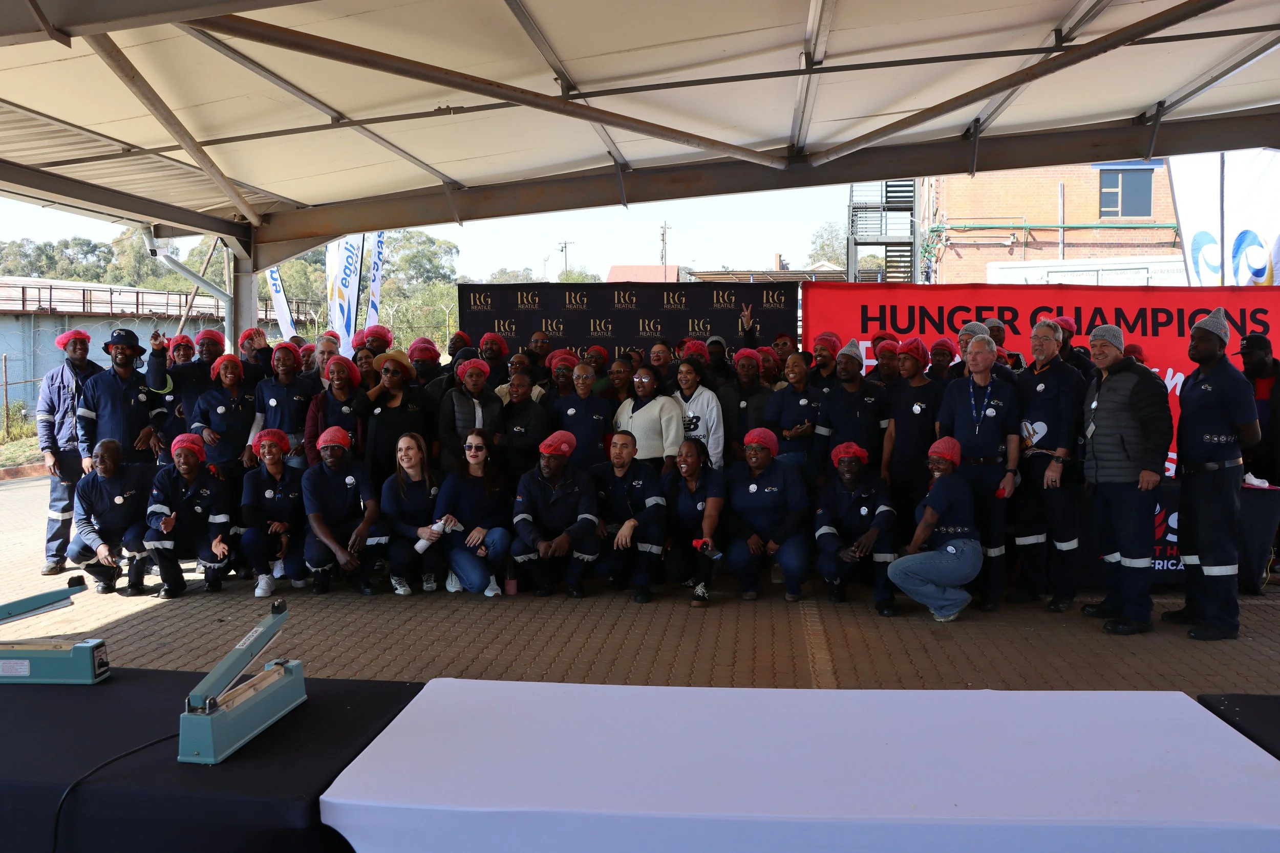 Group of people gathered under a large canopy at an outdoor event, with a red banner reading 'HUNGER CHAMPIONS' in the background. Many are wearing matching navy blue shirts and red hairnets, smiling and posing for a group photo.