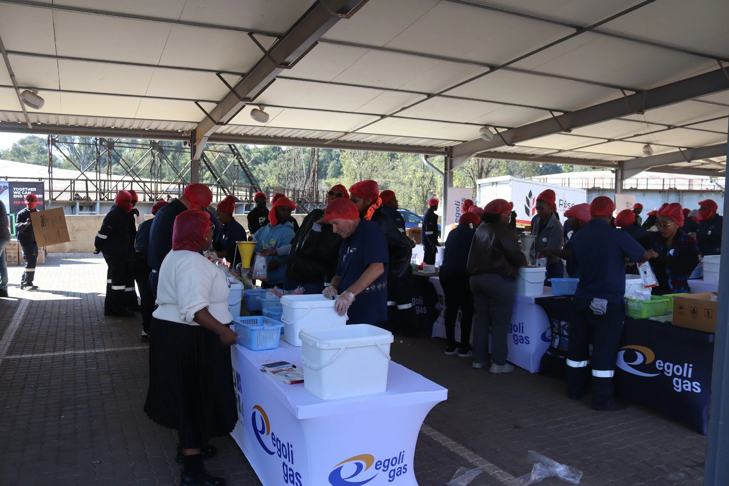 Group of people wearing red hairnets and dark clothing working at a long table covered with Egoli Gas branded tablecloths at an outdoor event under a large shelter. Some individuals are sorting or packaging items, and there are cardboard boxes and pl