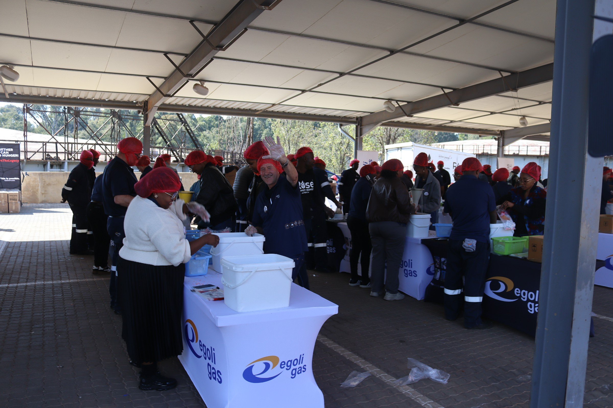 People wearing red hair coverings and work uniforms, serving and organizing food or supplies at an outdoor event under a large shelter, with banners and tables.