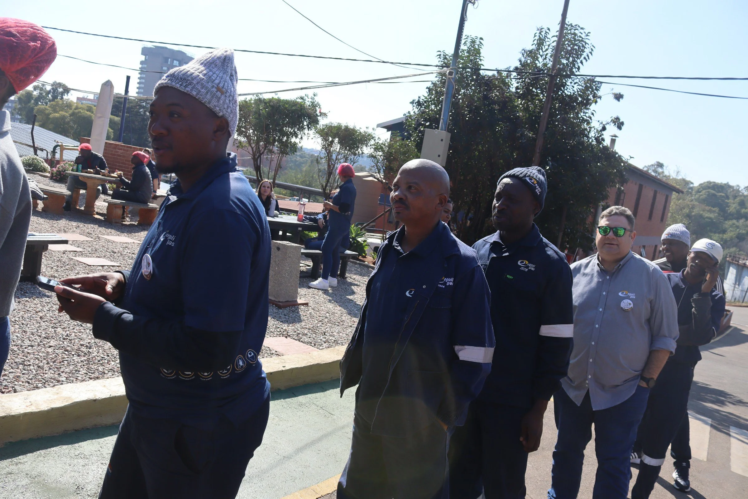 A group of people walking outdoors on a sunny day, some wearing jackets and beanies, with trees, buildings, and power lines in the background.