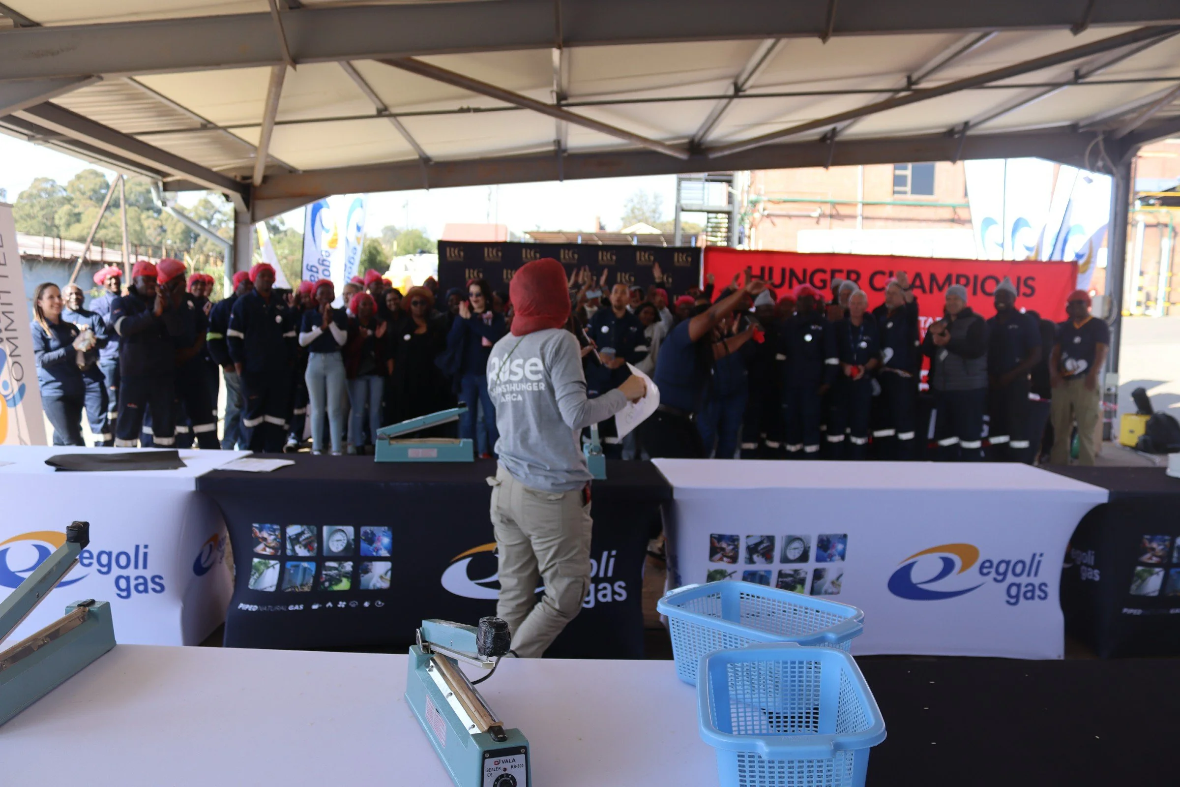 A large group of people, some wearing red head coverings, is gathered under a tent with a red banner that reads 'HUNGER CHAMPIONS'. A woman in a gray shirt stands in front of the group, gesturing as she speaks. In the foreground, there are tables wit