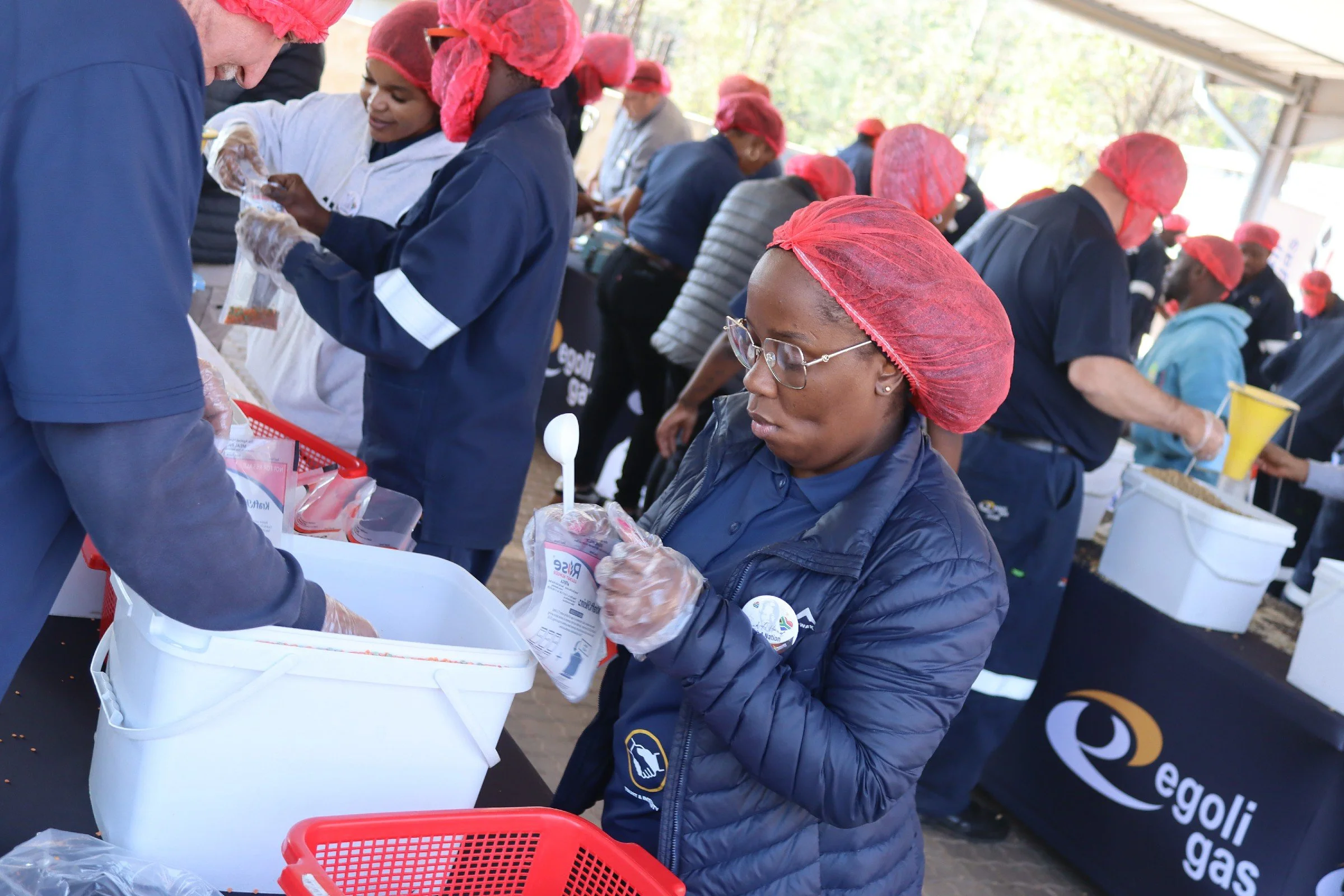 Group of people working together at a community service event, wearing red hair nets, with some packing items into white boxes outdoors.