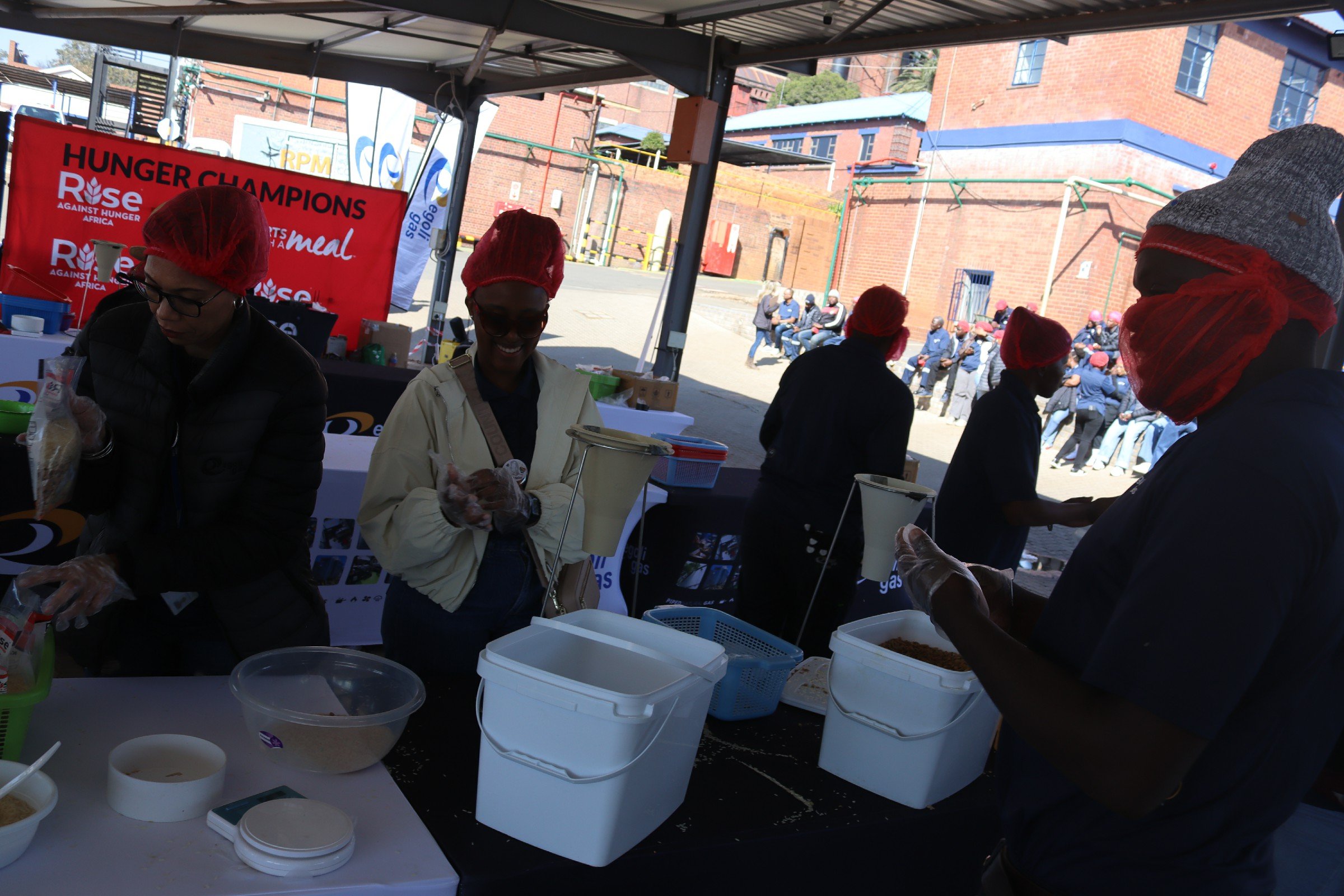 People wearing red hairnets and gloves working at a food distribution table under a tent during a community event, with a red banner that reads 'Hunger Champions' and 'Rise Against Hunger Africa' in the background, and a line of people waiting outsid