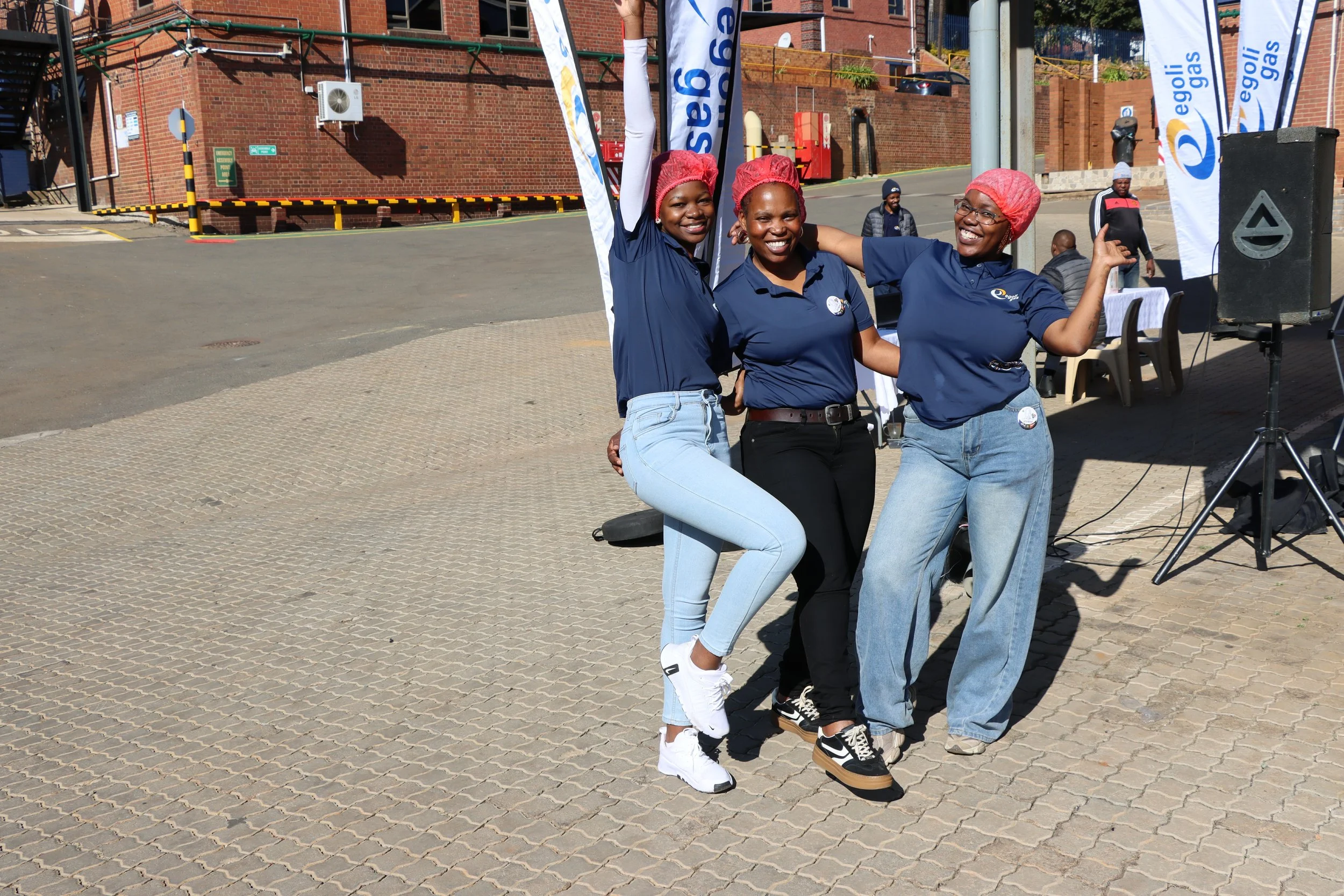 Three women wearing blue shirts, jeans, and red hair nets are smiling and posing together outdoors near a charity event setup with banners and equipment. One woman is giving a thumbs-up, and they are standing on a paved area with a brick building in 