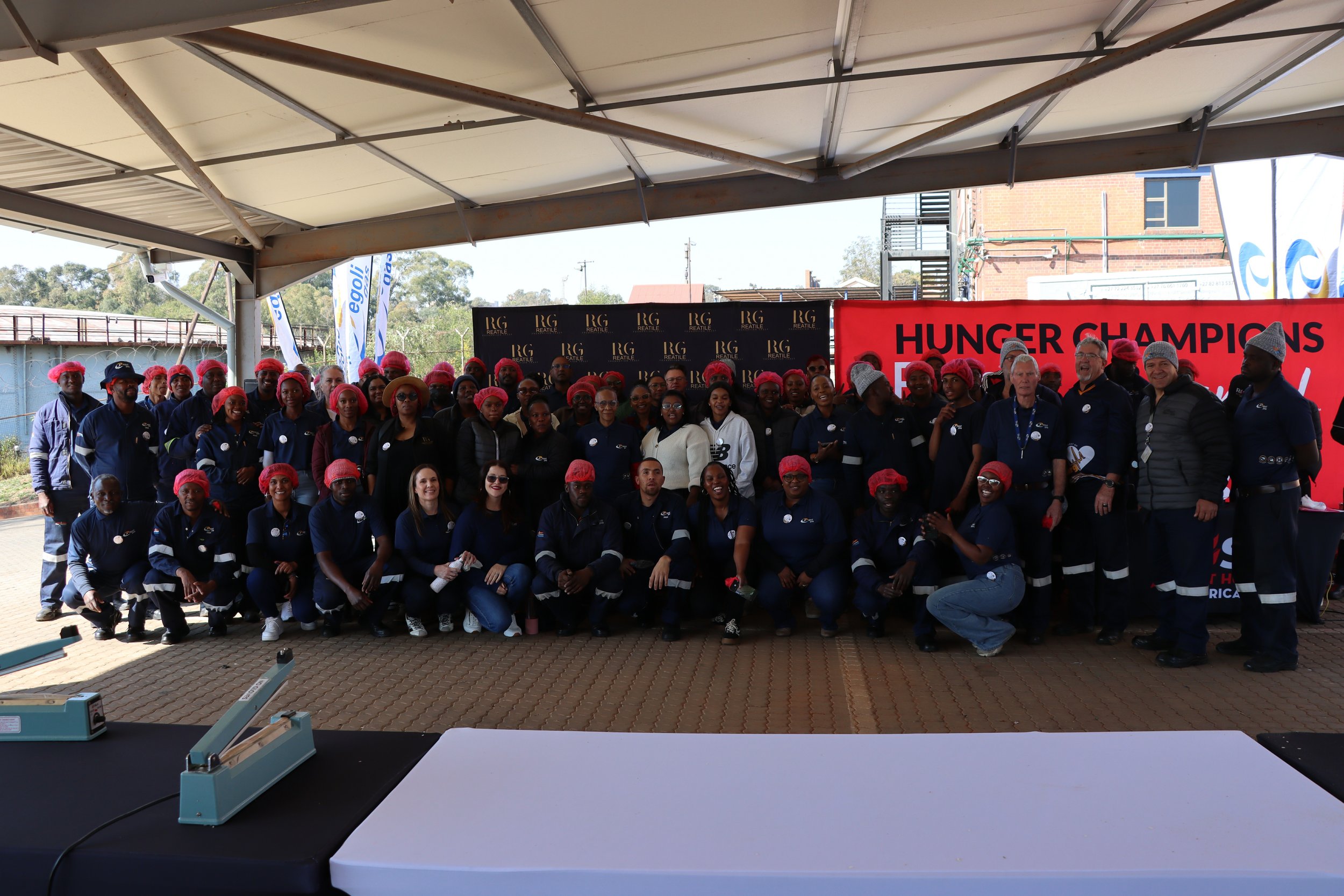 A large group of people, many wearing pink hairnets and dark uniforms, gathered under a canopy in front of a red banner that reads 'HUNGER CHAMPIONS'. The group includes men and women of various ages and ethnicities. The background shows a brick buil