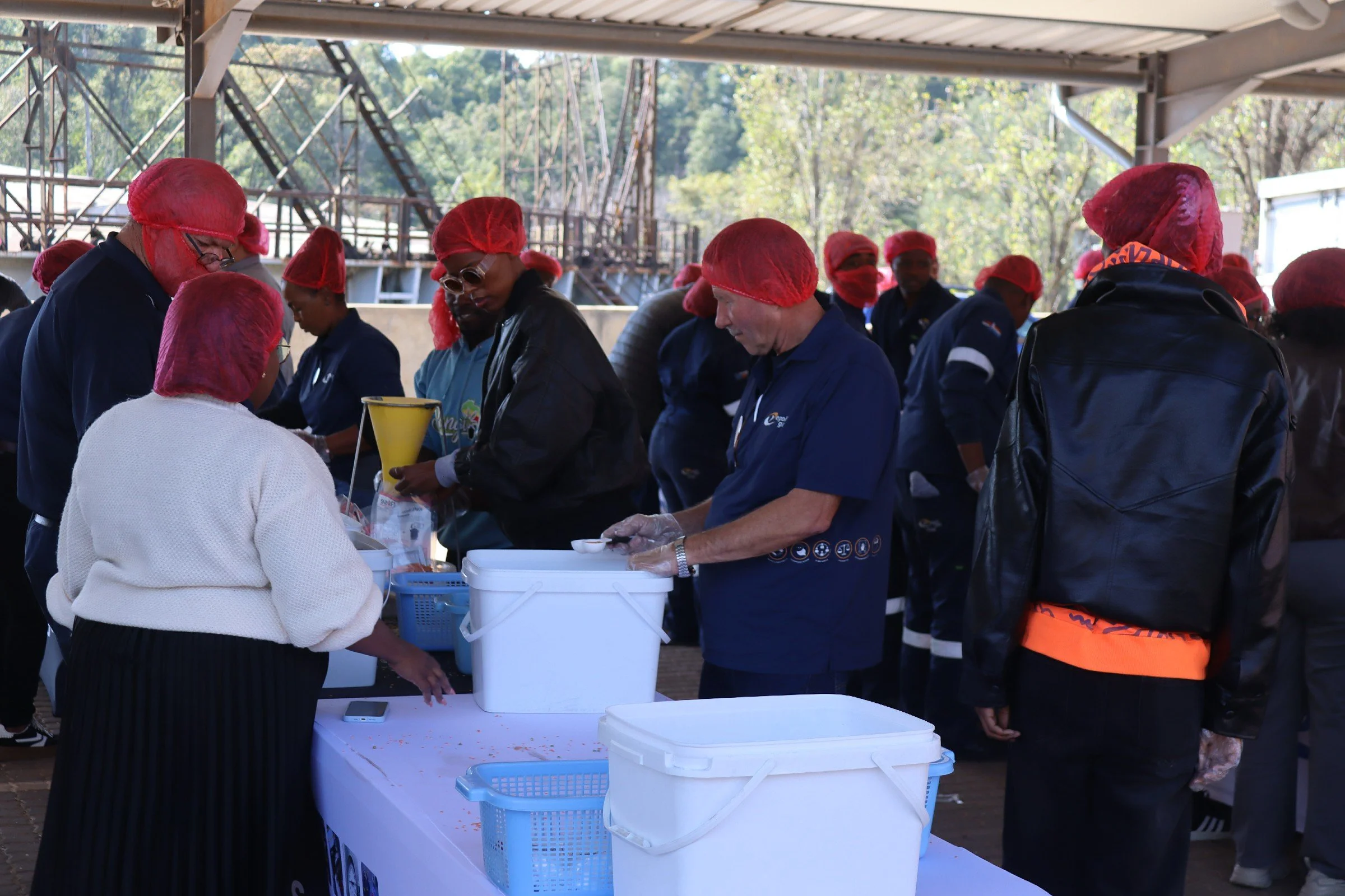 People wearing red hairnets and blue uniforms standing around a table outdoors, serving or preparing food under a covered area with trees in the background.