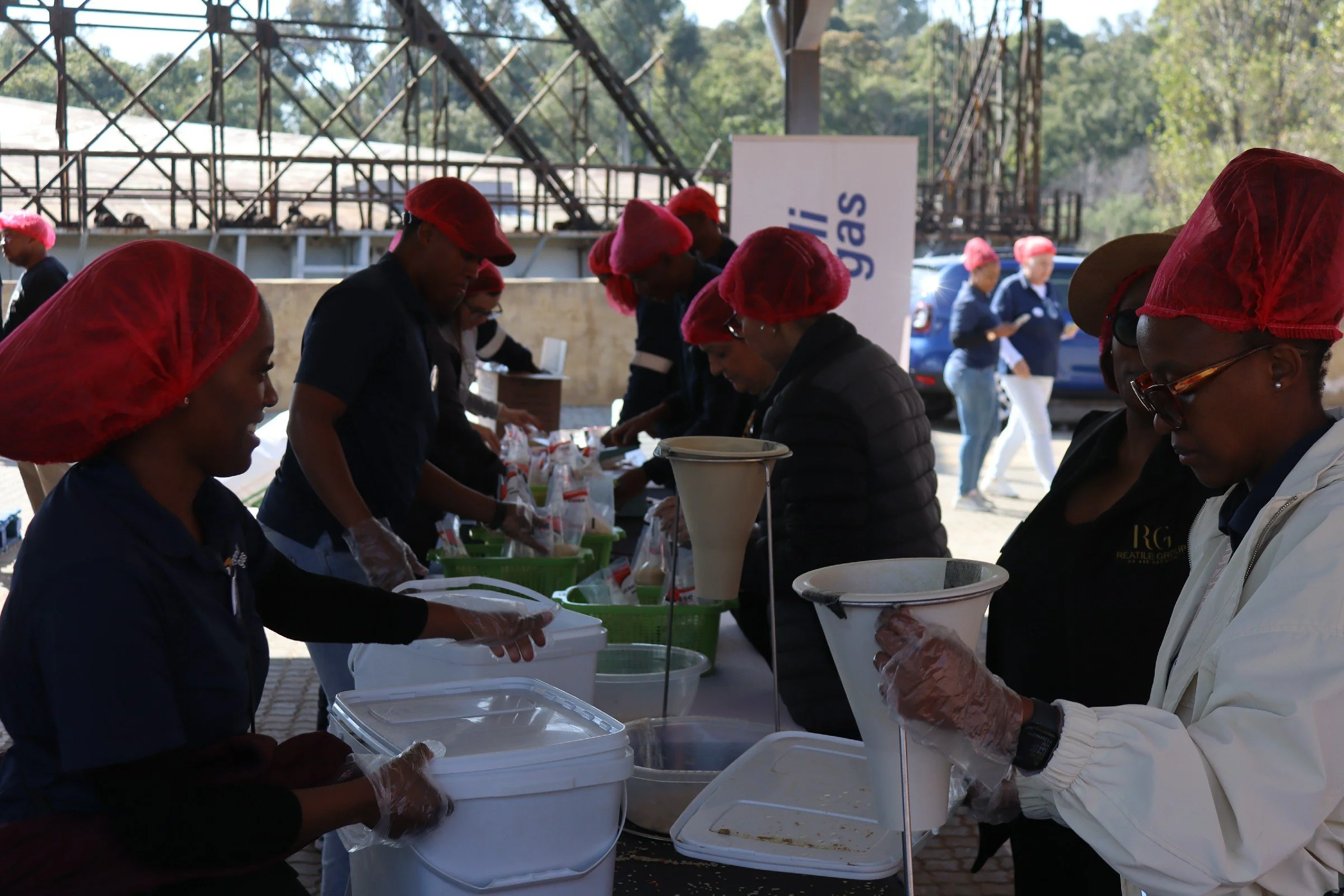 People wearing red hairnets and gloves serving food at an outdoor event with tables and white containers, surrounded by trees and metal structures.