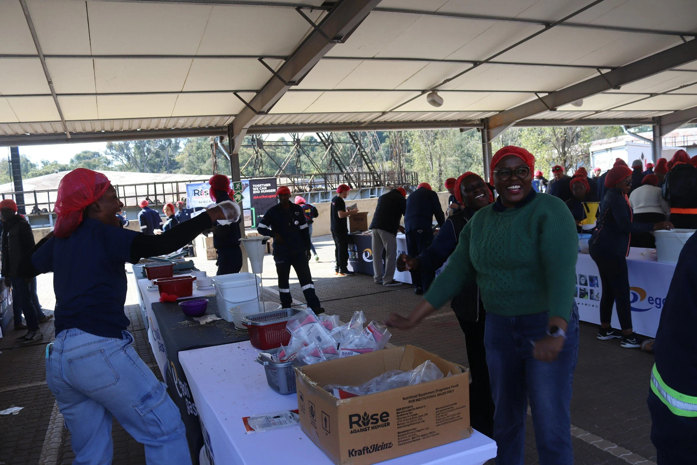 People wearing red hair covers at a food distribution event, with tables filled with food packages and a sign that reads 'Raise Against Hunger'.