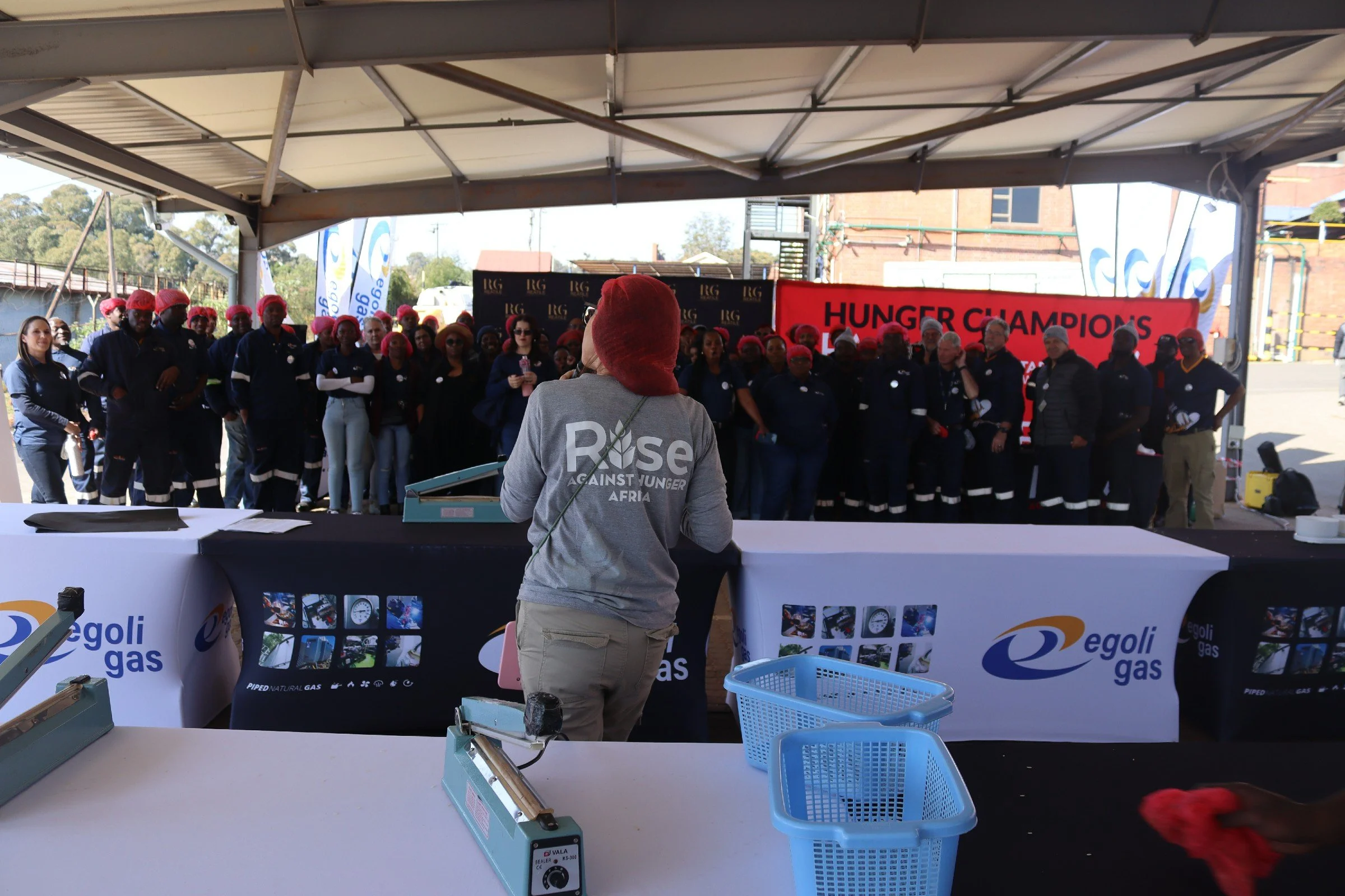 A group of people wearing red hairnets and dark uniforms standing under a covered outdoor structure during daytime, with a woman in the foreground wearing a grey shirt with 'Rise Against Hunger Africa' written on the back. There is a large red banner