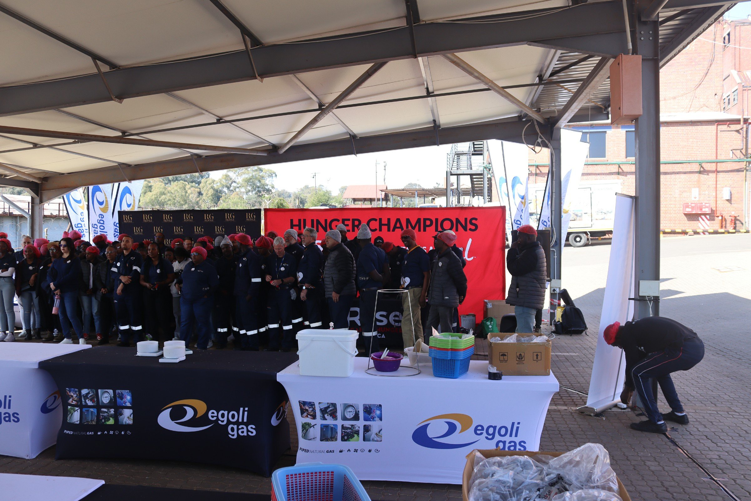 Outdoor event under a tent with a group of people, many wearing red hats, standing in front of a red banner that reads 'HUNGER CHAMPIONS.' There are tables with supplies and Egoli Gas branding visible, and a person is taking photos or recording near 