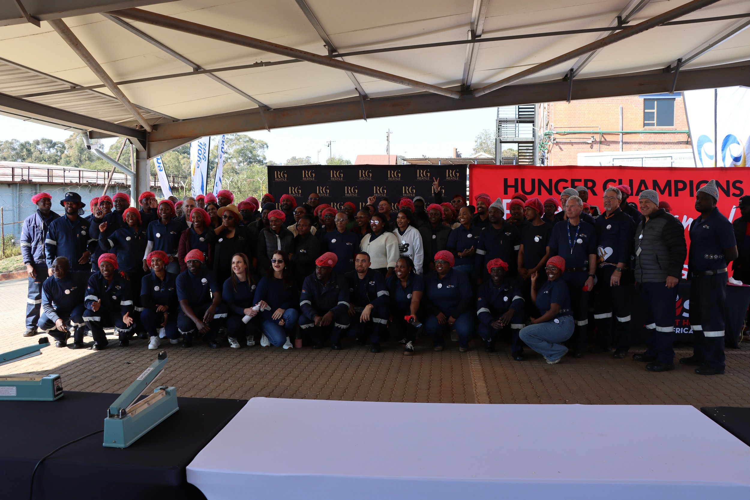 Group of diverse people wearing red hairnets and navy uniforms at an outdoor event with banners and a stage.