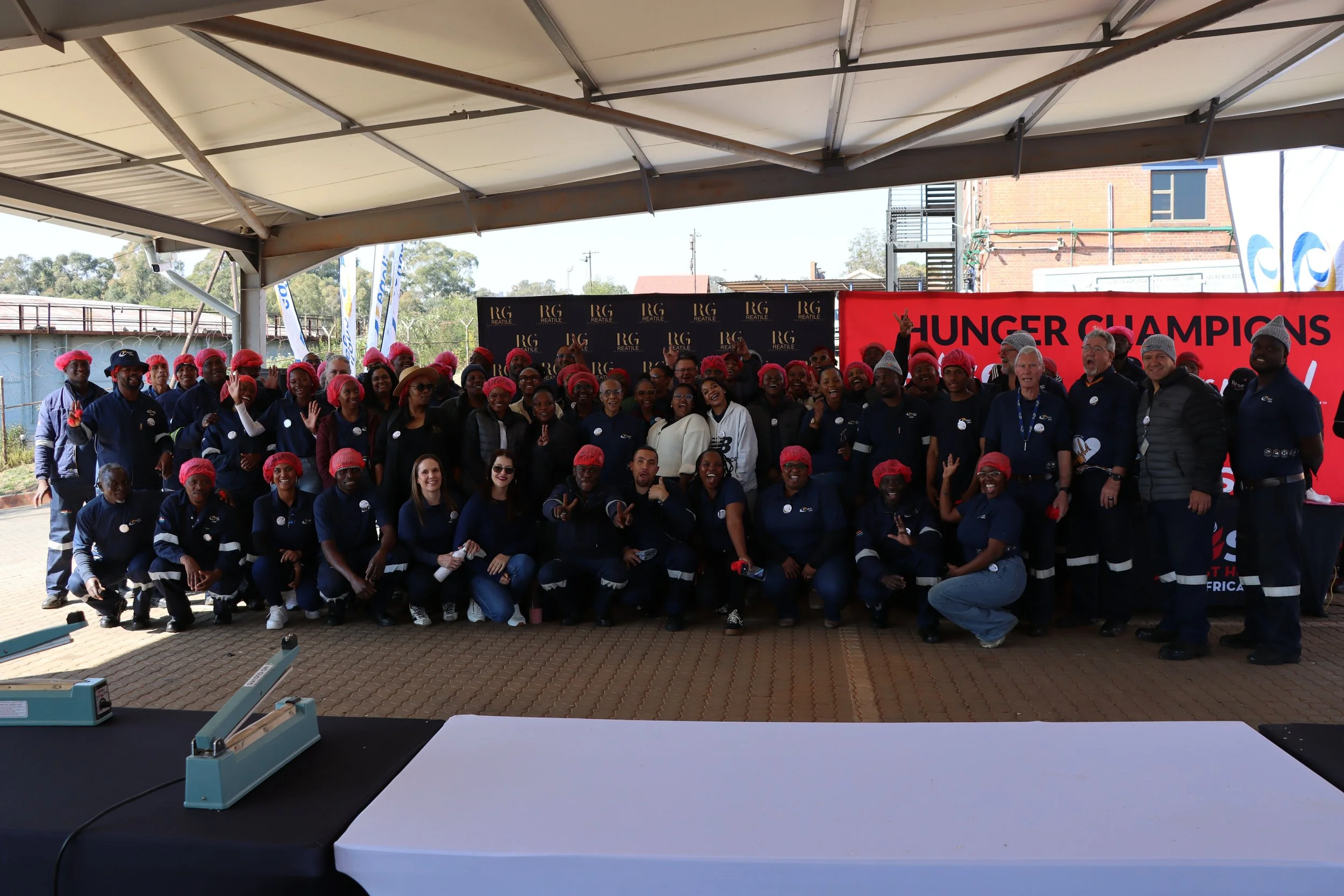 Group of people posing for a photo in front of a banner that says 'HUNGER CHAMPIONS'. They are under a large tent, some wearing pink hairnets, in an outdoor setting. There is a table with paper and a machine in the foreground.