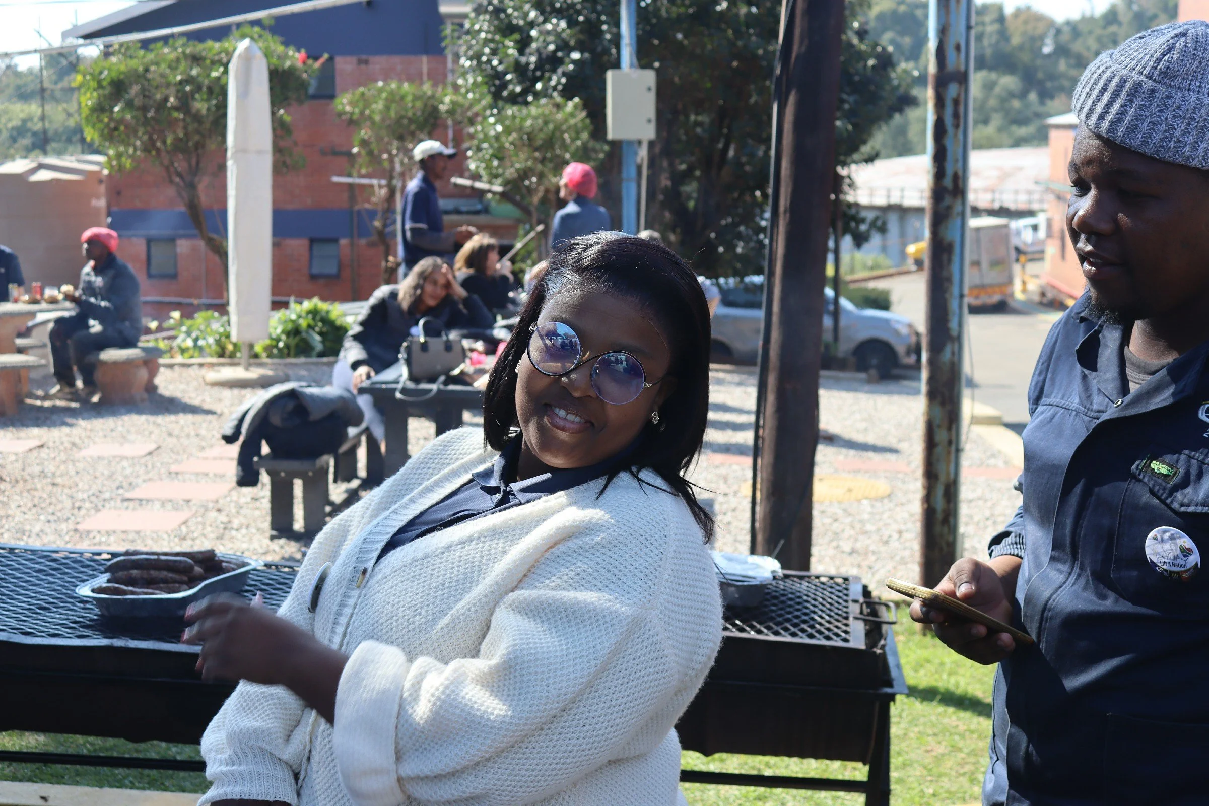 Two women standing outdoors, one smiling and wearing glasses, the other looking down at her phone, with people in the background near a grill, trees, and a brick building.