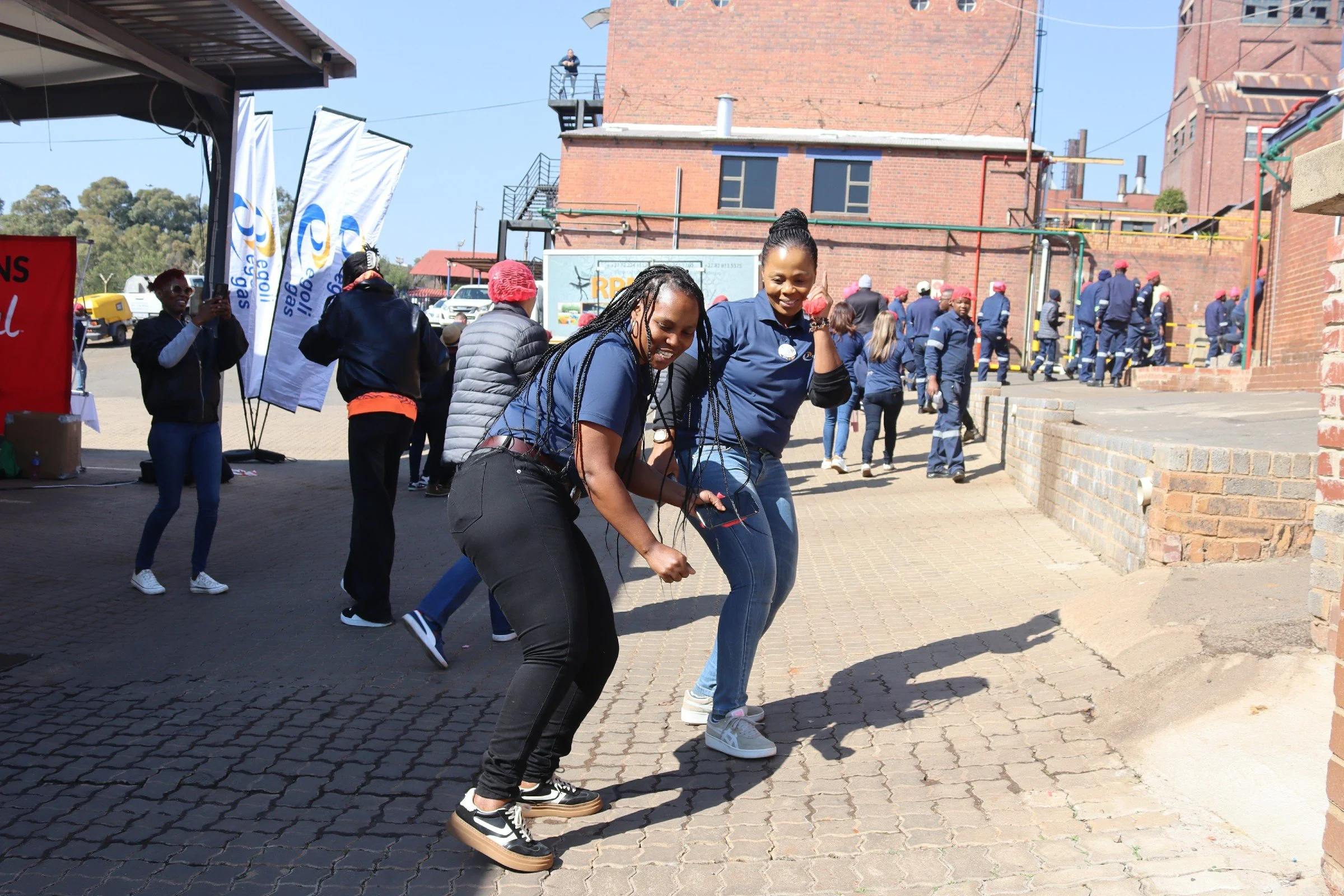 Two women in blue shirts engaging in a jumping or dancing activity outdoors at a public event, with a group of people and a brick building in the background.