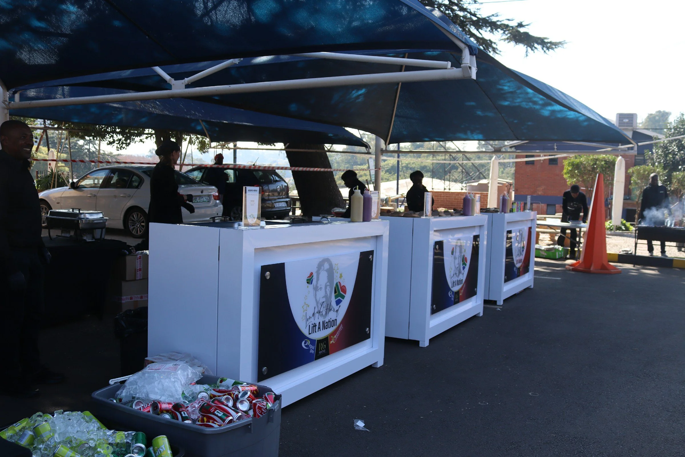 Outdoor event booth with tables and banners, cans and bottles on the ground, and people standing around, under large blue canopies. Cars are parked behind the booth, and trees and buildings are in the background.