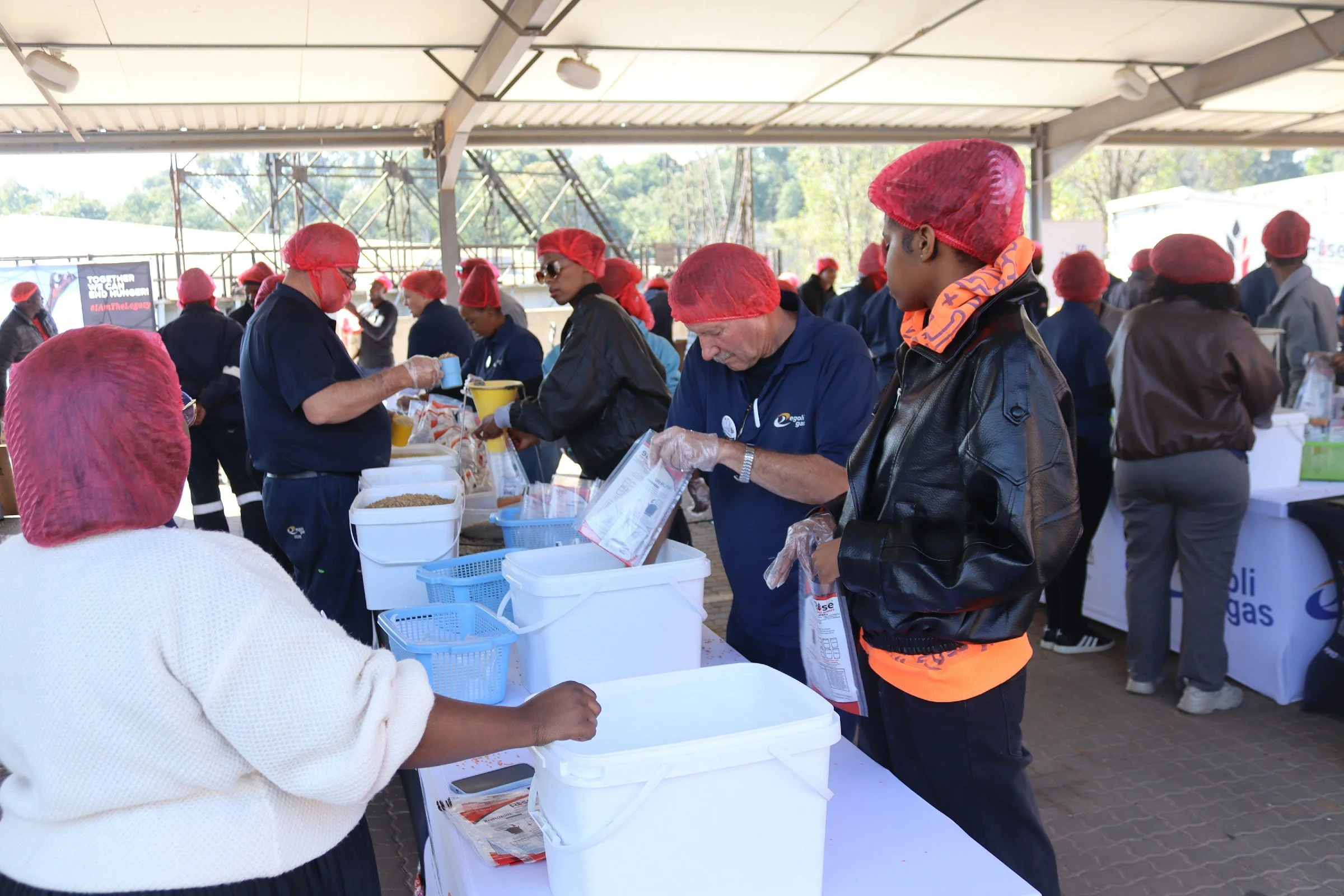 People wearing red hair nets and gloves preparing and distributing food at a community event or charity drive under a large outdoor shelter.
