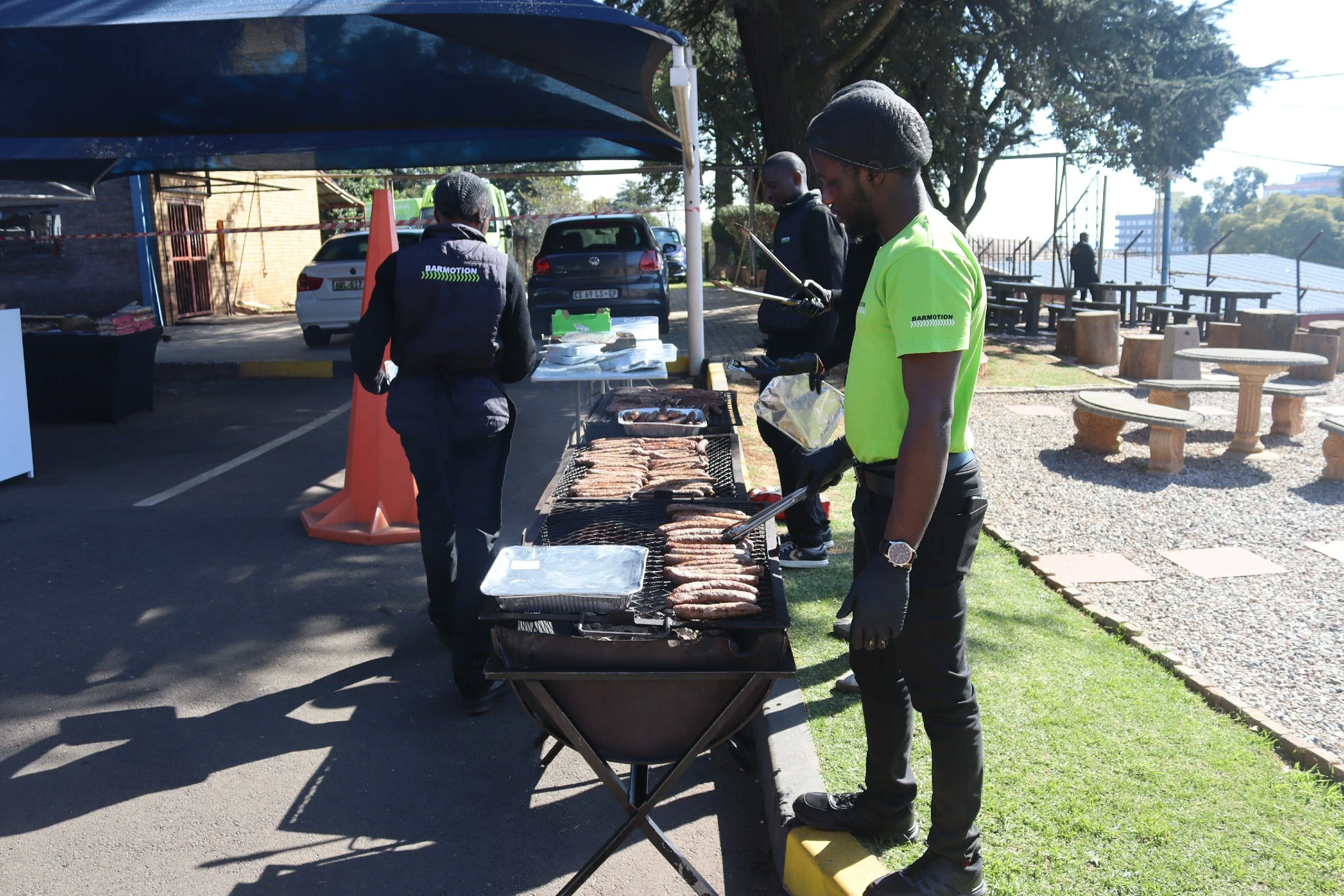 People grilling and cooking meat at an outdoor barbecue event under a canopy, with parked cars and trees in the background.