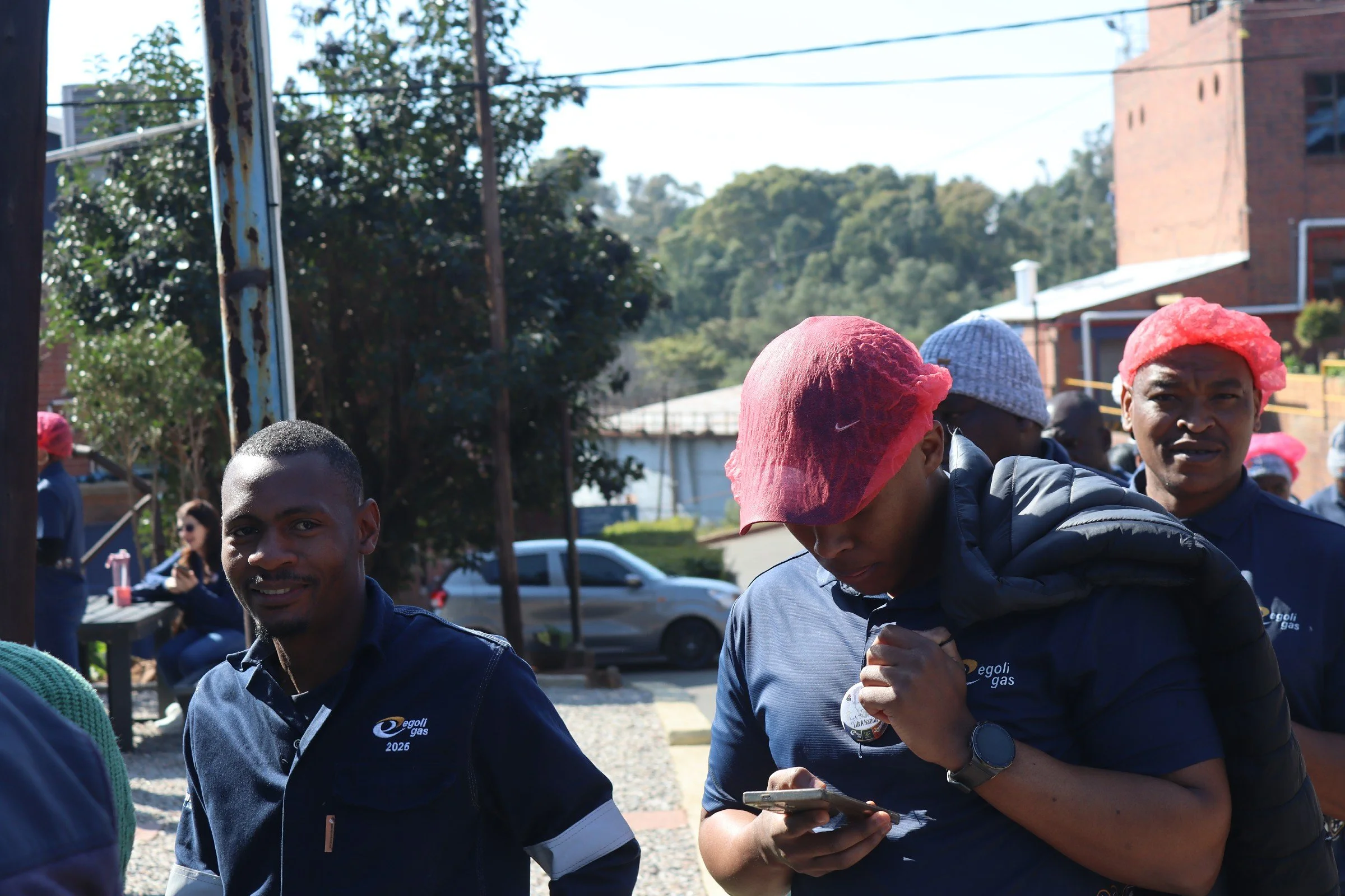 A group of people wearing matching navy blue jackets with a logo, some with pink hairnets, gathered outdoors on a sunny day, with trees, cars, and buildings in the background.