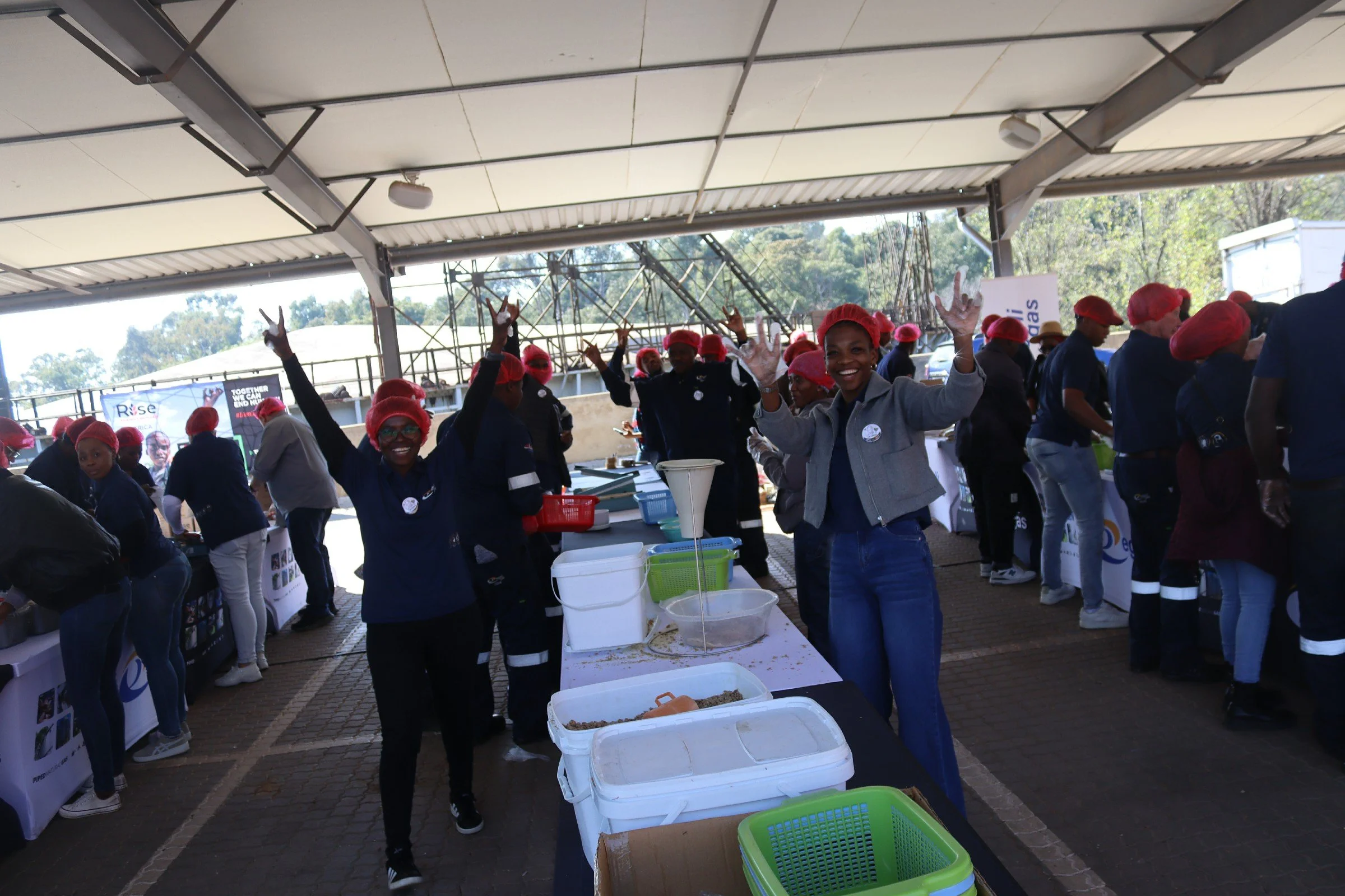 A group of people wearing red hair coverings and gloves, smiling and waving in an outdoor market or event setting under a large canopy. There are tables with containers and bottled drinks.