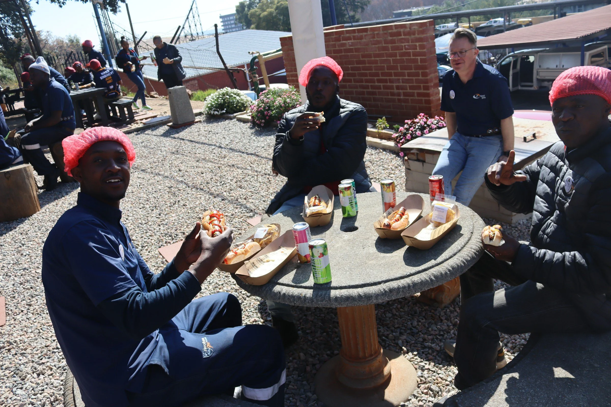 People enjoying a meal outdoors at a round stone table, with some holding hot dogs, drinks, and giving thumbs up, under sunny weather with a background of a brick wall, flowers, and parked cars.