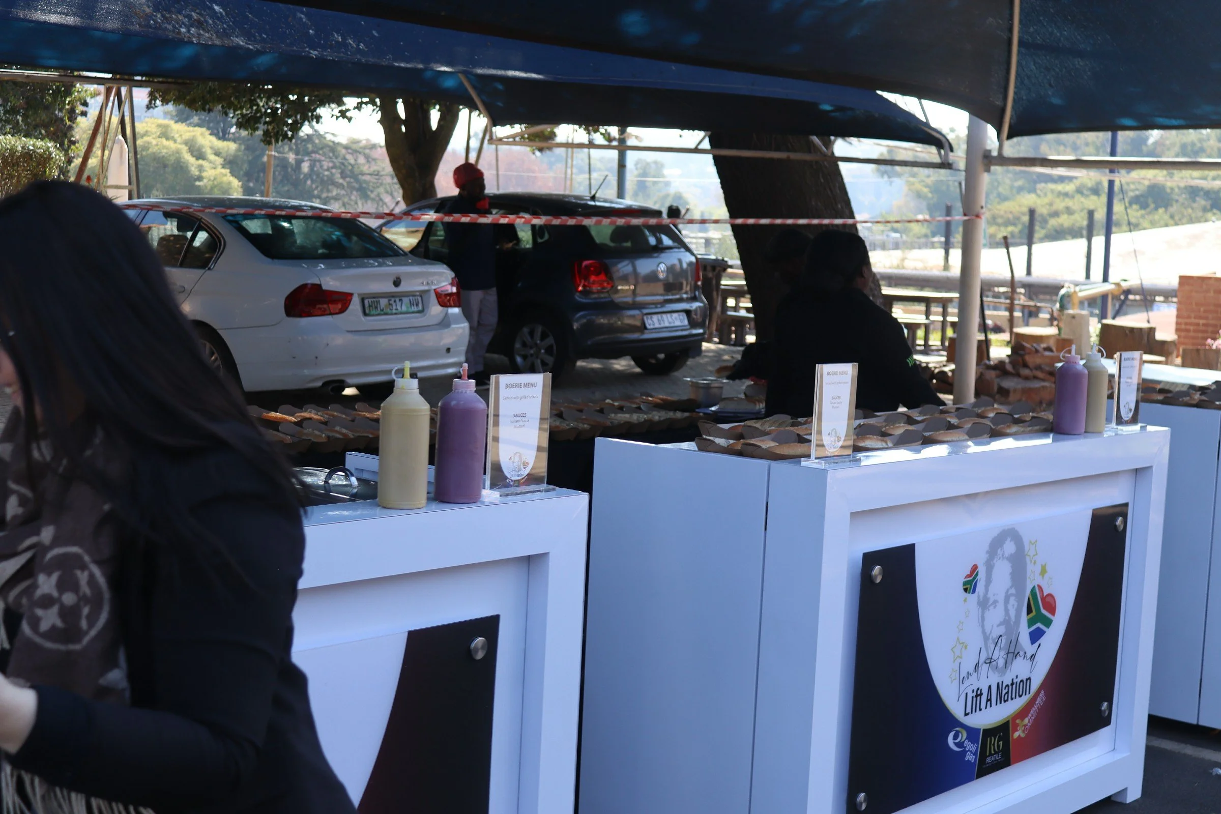 Food stand with condiments and signs, cars parked behind, and people around at an outdoor event.