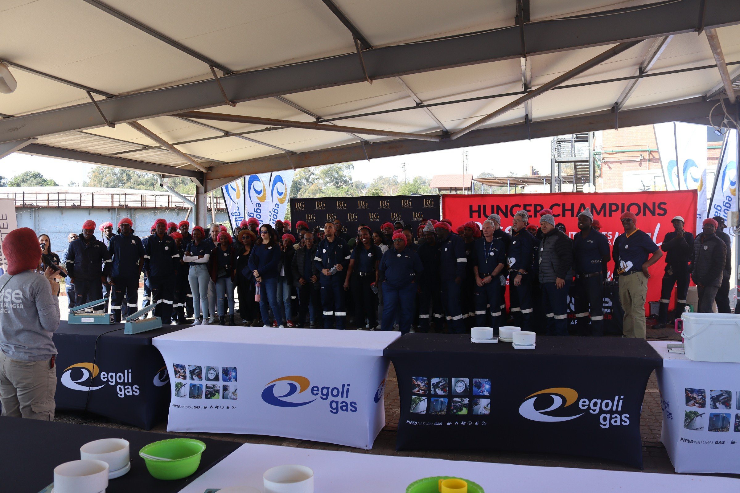 Group of people wearing dark uniforms and red hats standing under a large canopy at an outdoor event, with banners and tables displaying logos and promotional materials for Egoli Gas, during a hunger relief event.