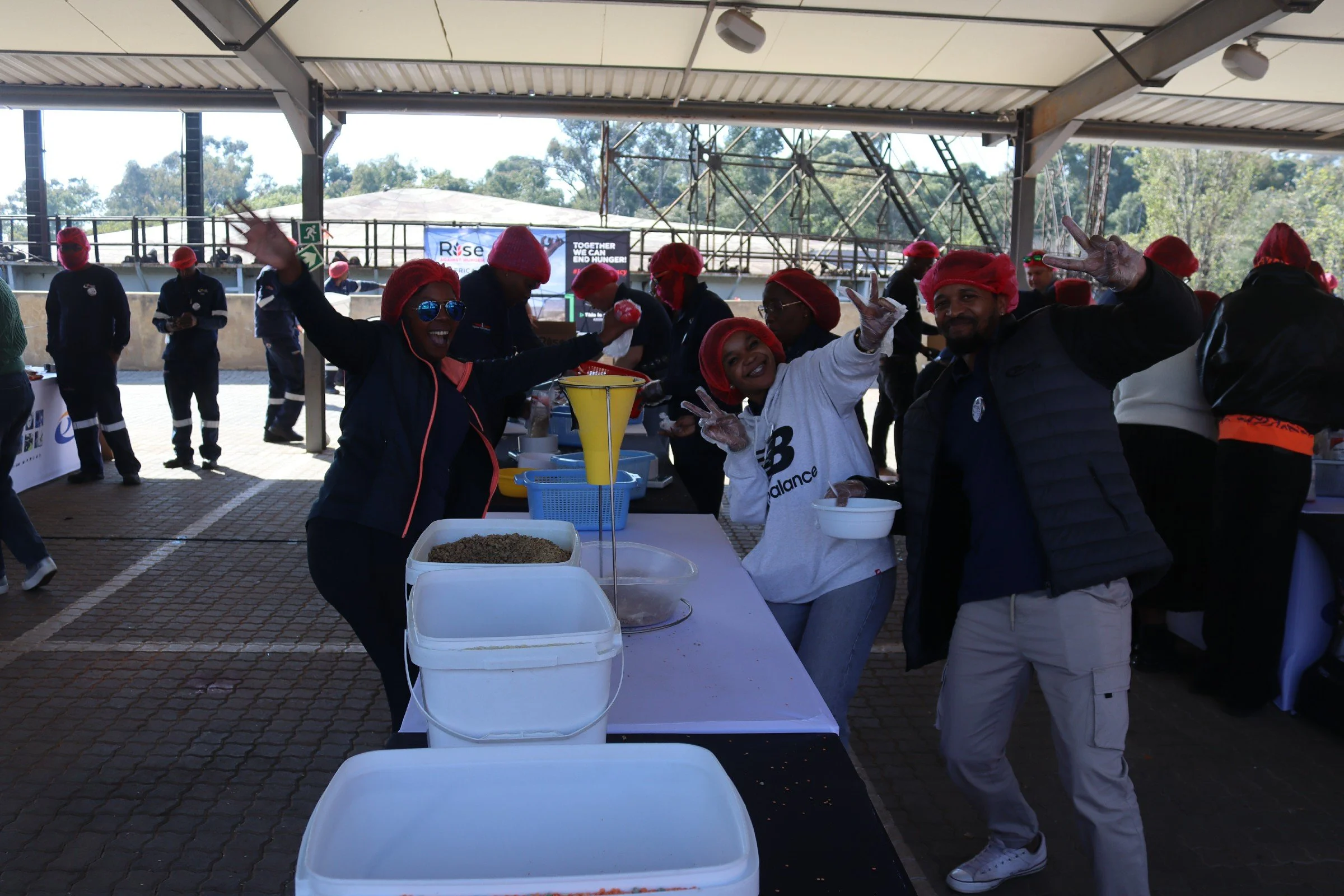 People wearing red head coverings and gloves standing and smiling at a food distribution event under a covered outdoor area. There are containers of food on tables and some people in the background.